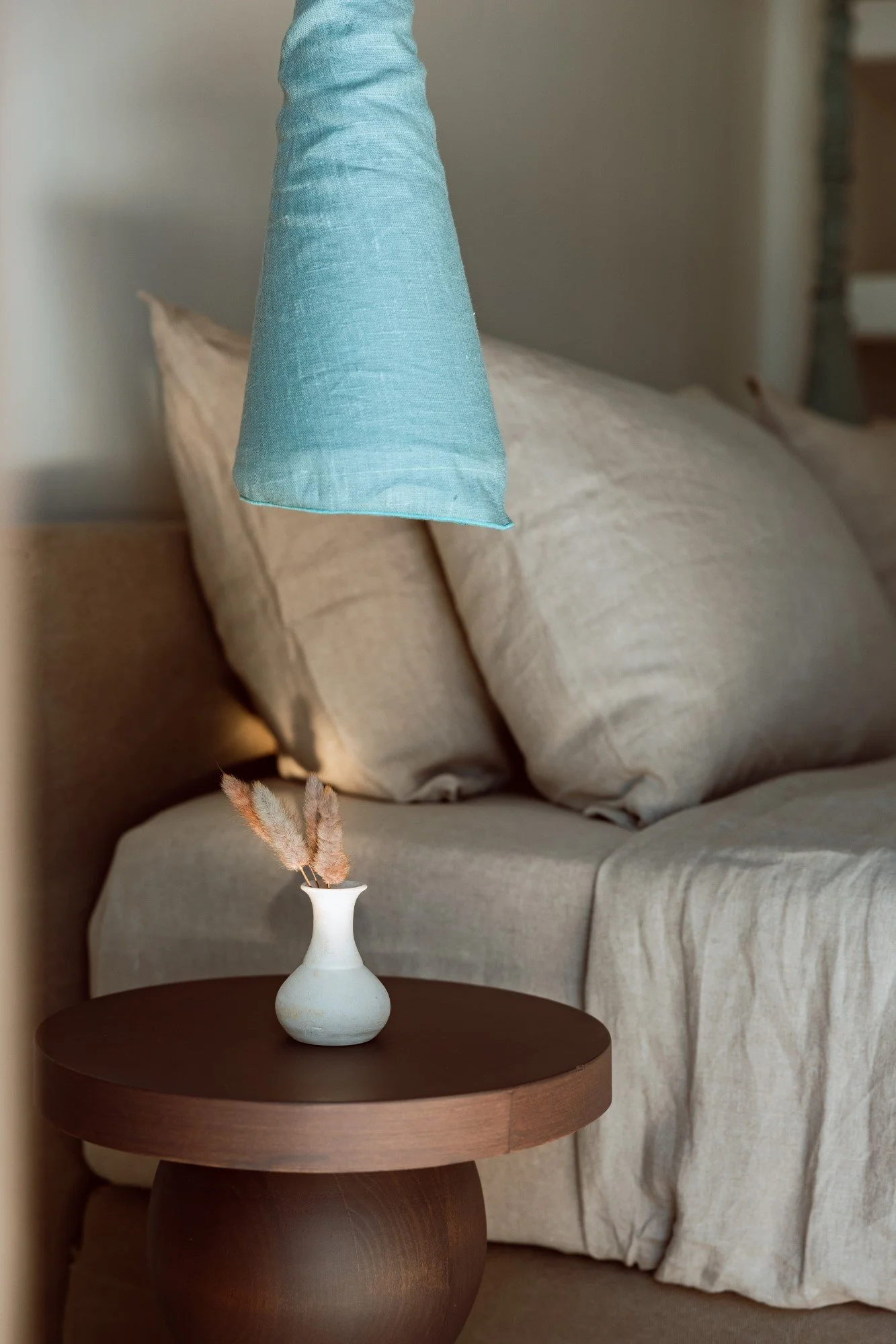 A cozy living room corner with a beige sofa, beige pillows, a small round wooden side table, a white vase with dried grasses, and a teal lampshade hanging above.