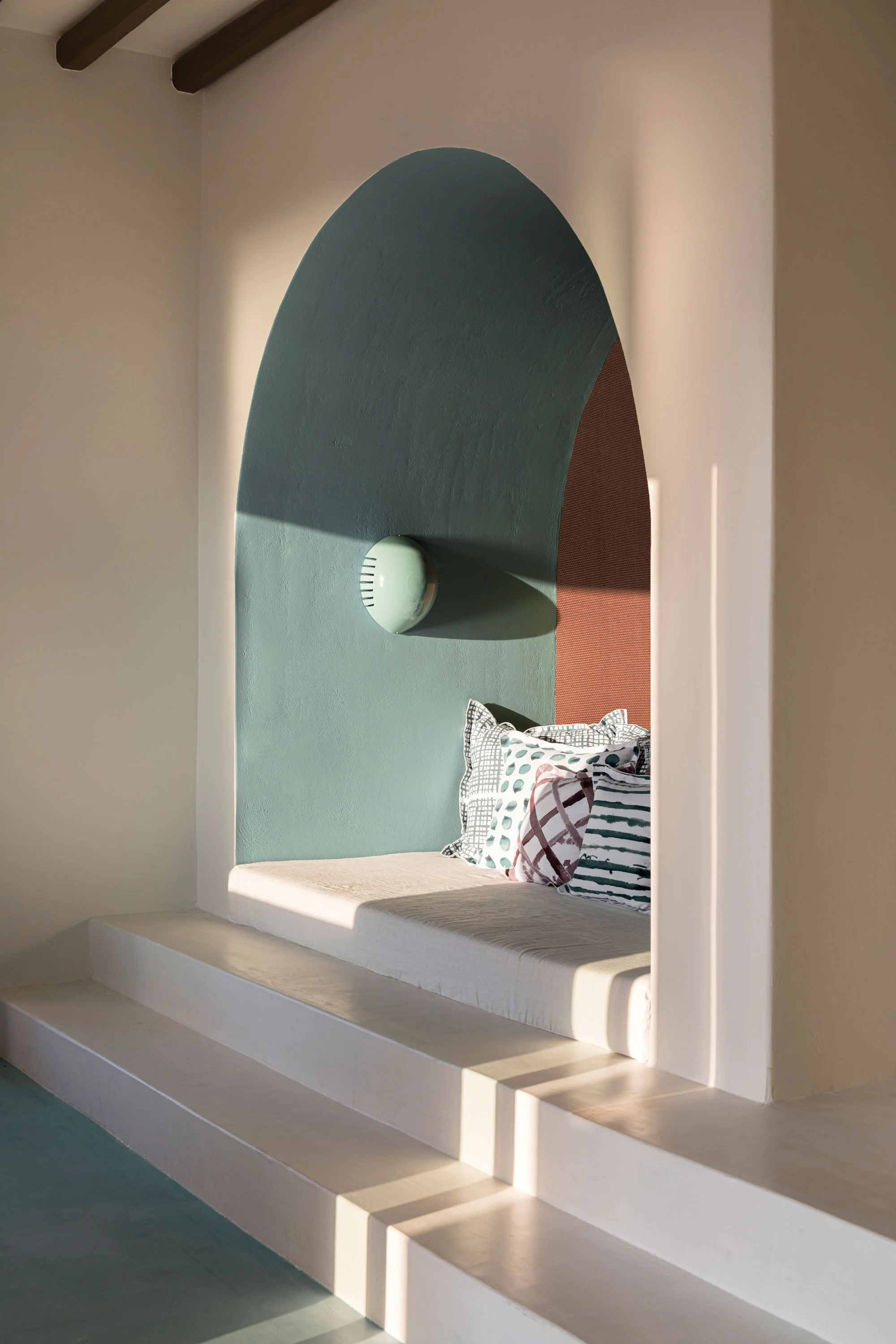 A built-in seating nook with steps leading up to a cushioned seat decorated with striped and patterned pillows, set within an arched wall with pastel colors and modern lighting.