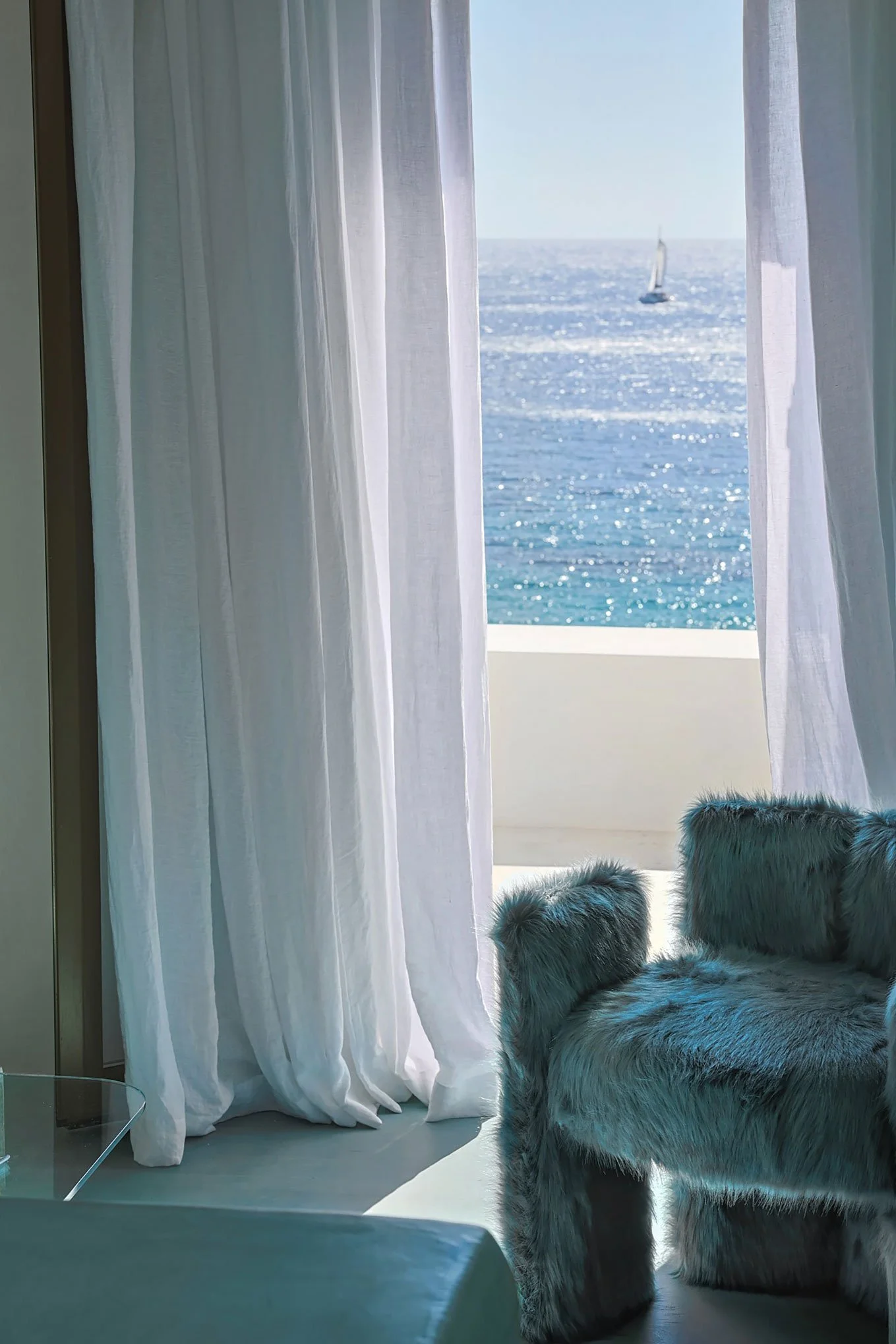 Room with white curtains overlooking the ocean, sailboat in the distance, fur-covered armchair in foreground.