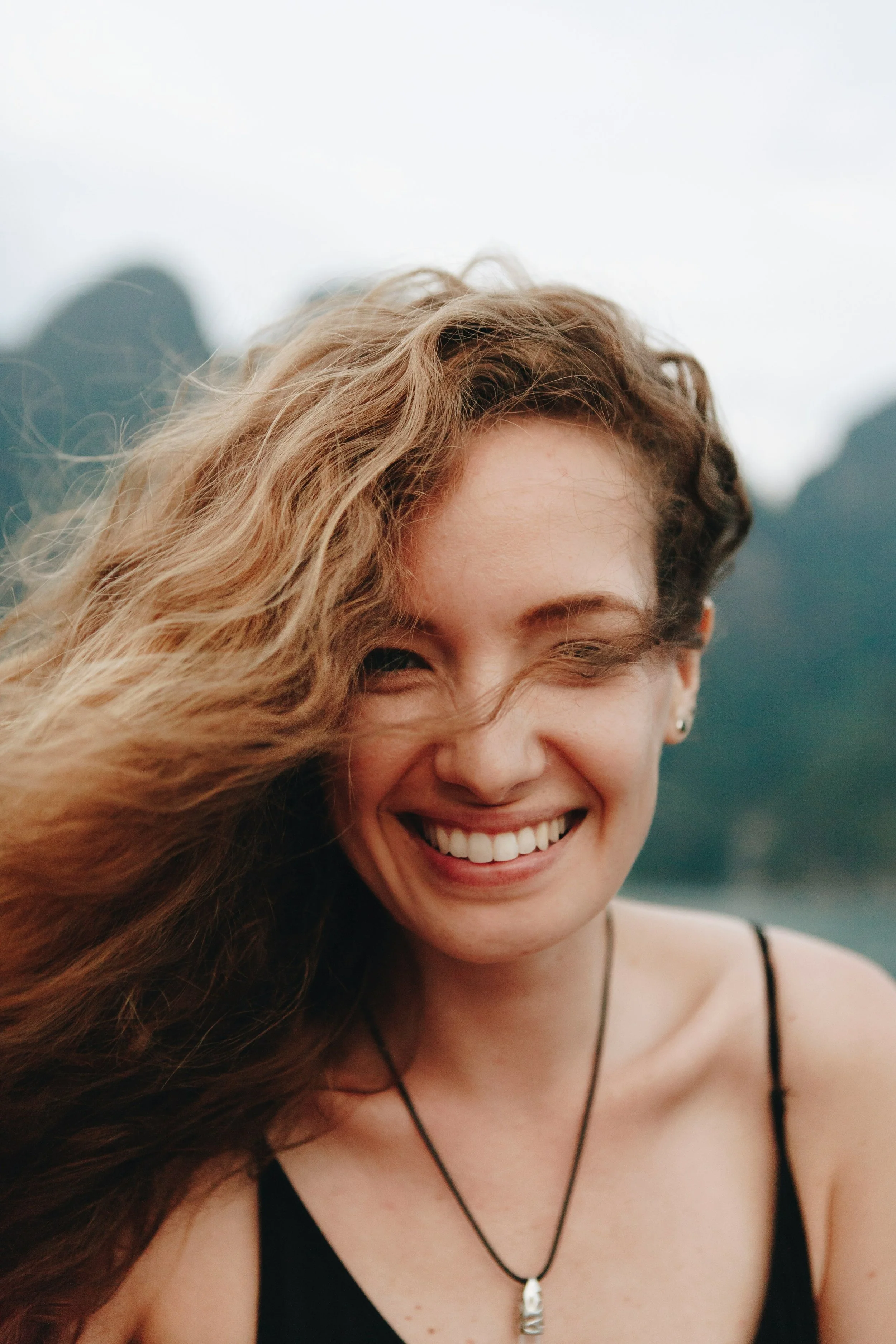 A young woman with curly blonde hair smiling outdoors, wearing a black spaghetti strap top and a necklace, with a blurred natural background.