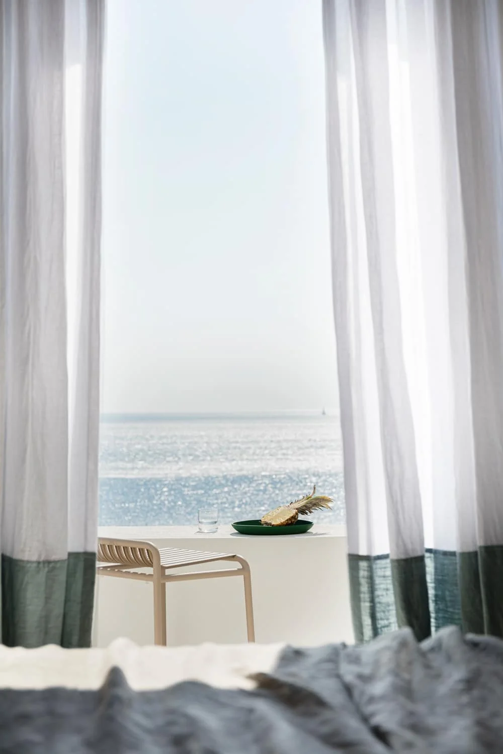 View of the ocean through open white curtains, a table with a glass of water and a plate with a piece of bread and a feather, as seen from inside a room.