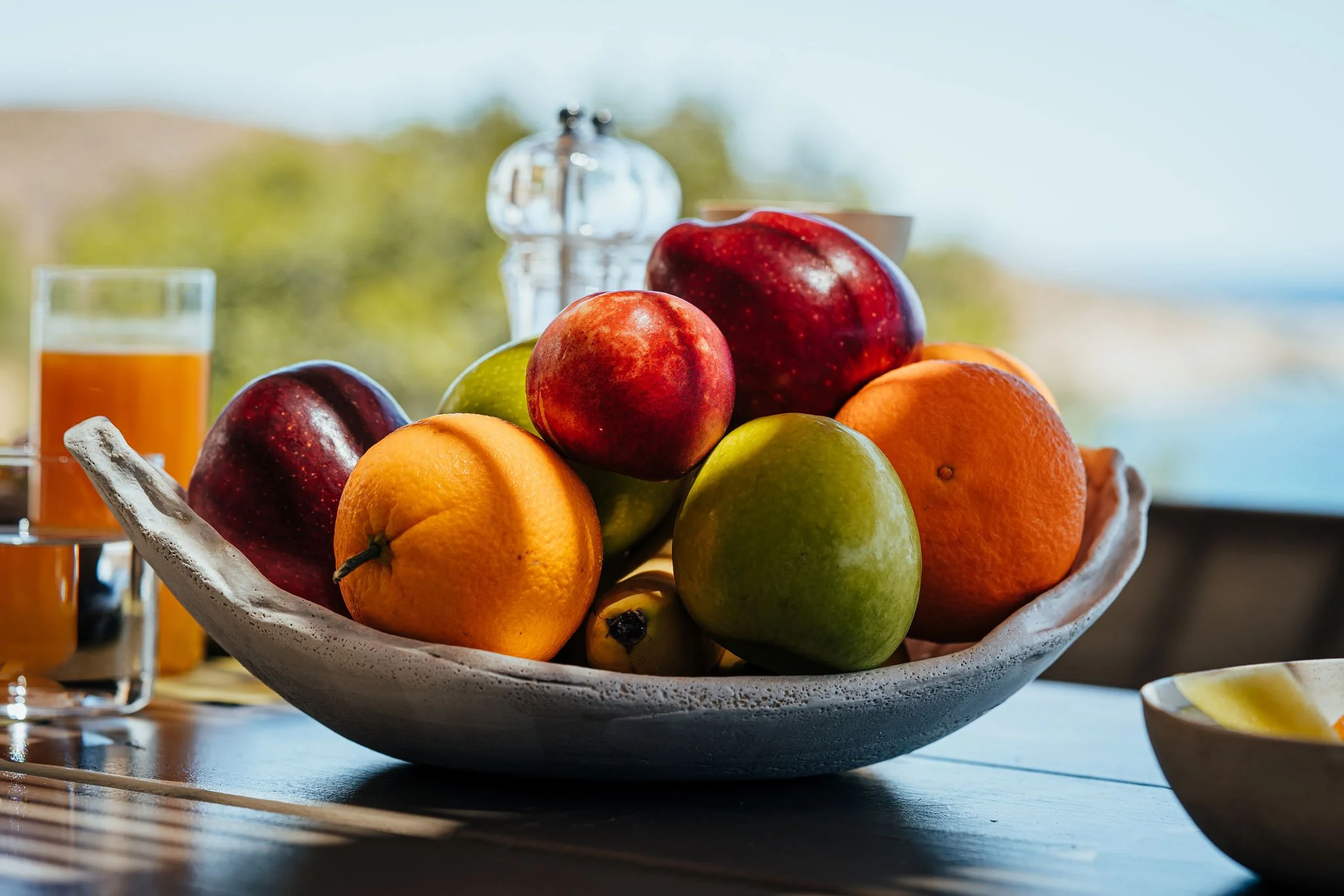A bowl of assorted fresh fruits including apples, oranges, bananas, and pears on a wooden table outside.