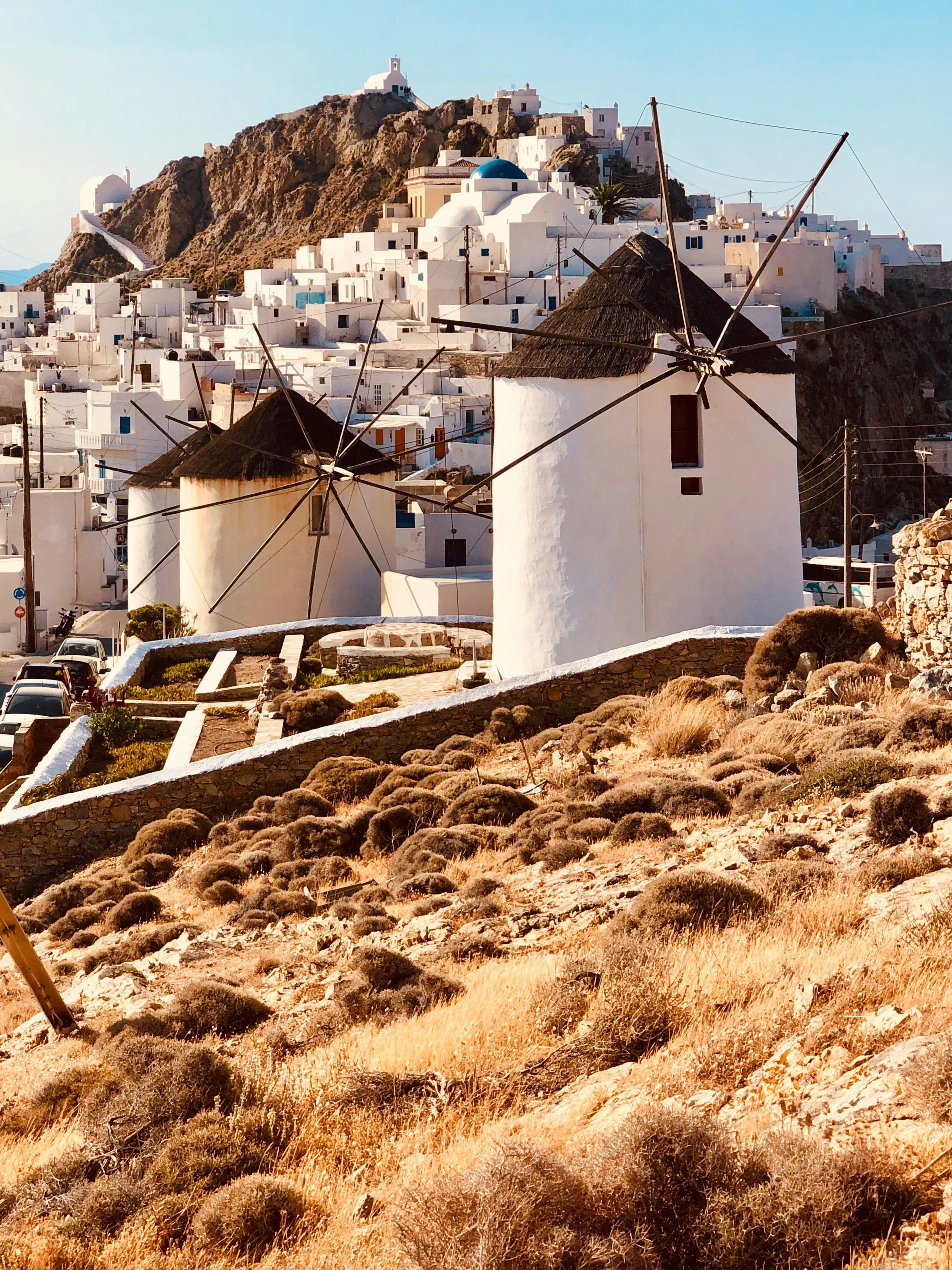 Two traditional windmills with dark thatched roofs in a dry, rocky landscape in front of white buildings on a hillside, typical of Santorini, Greece.