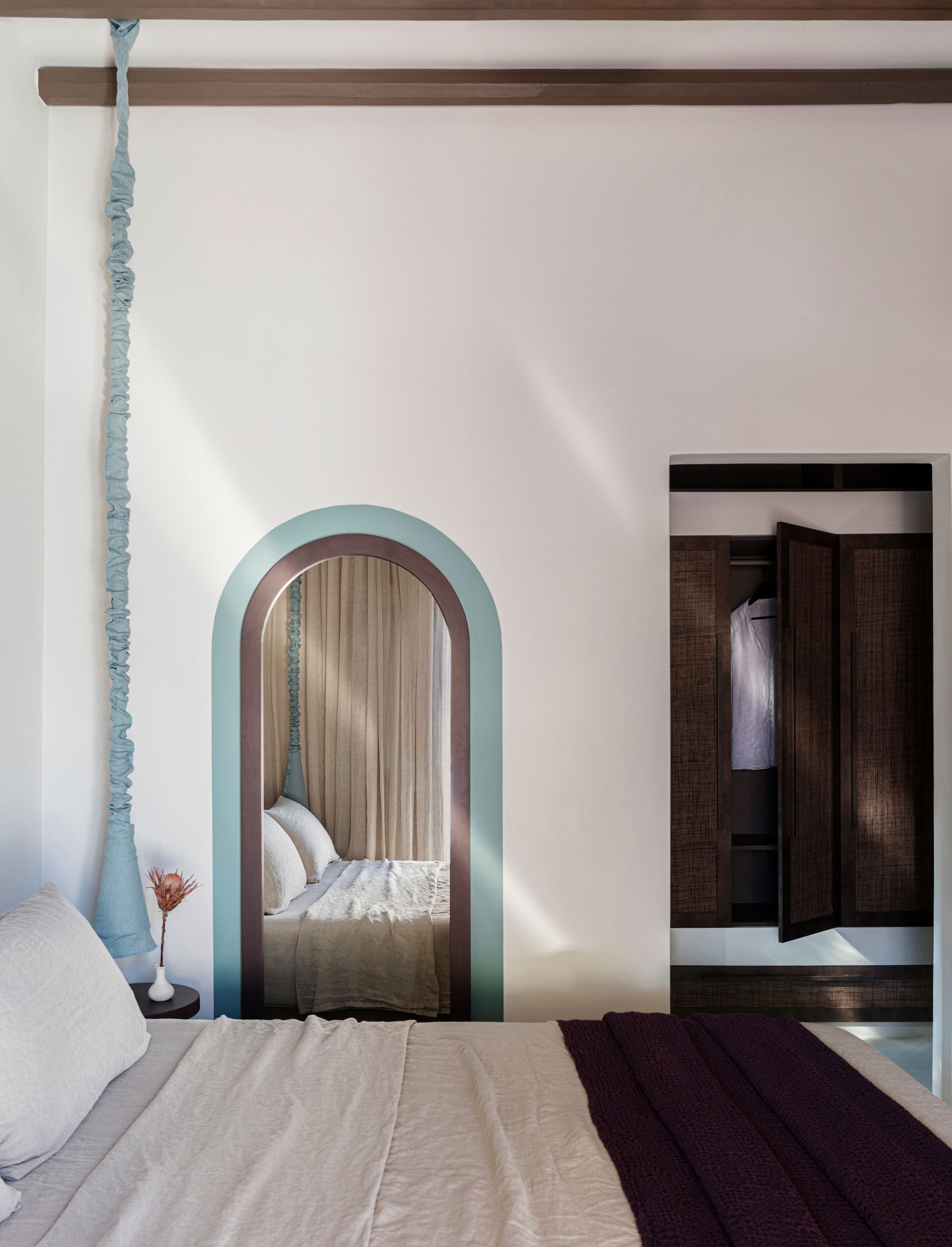 Bedroom with a bed, a mirror with an arched frame, a wardrobe with open doors, and a small side table with a vase and dried flowers.