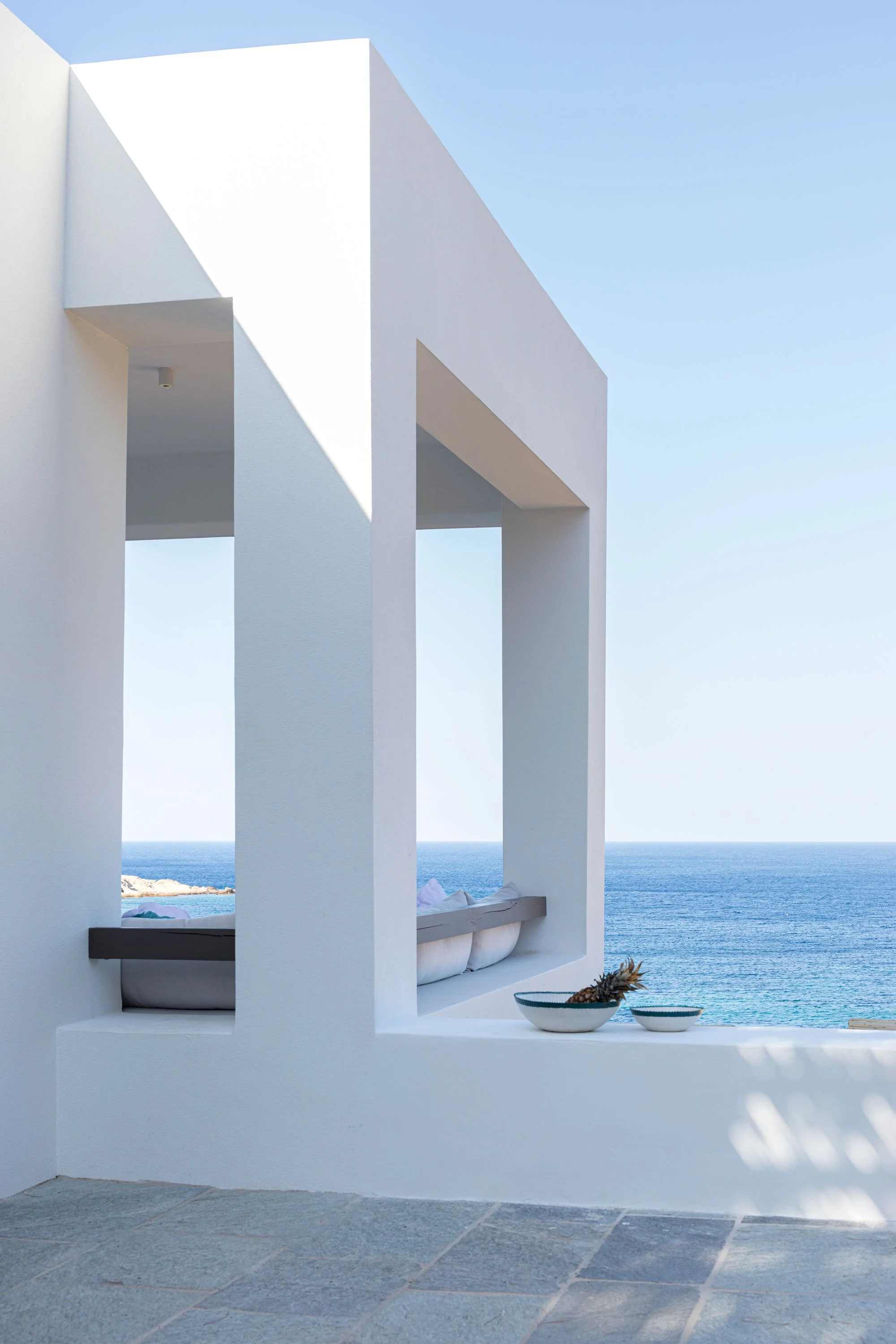 Modern white architectural structure with sea view, featuring open rectangular frames, a built-in bench with cushions, and bowls with a pineapple and fruit, under a clear blue sky.