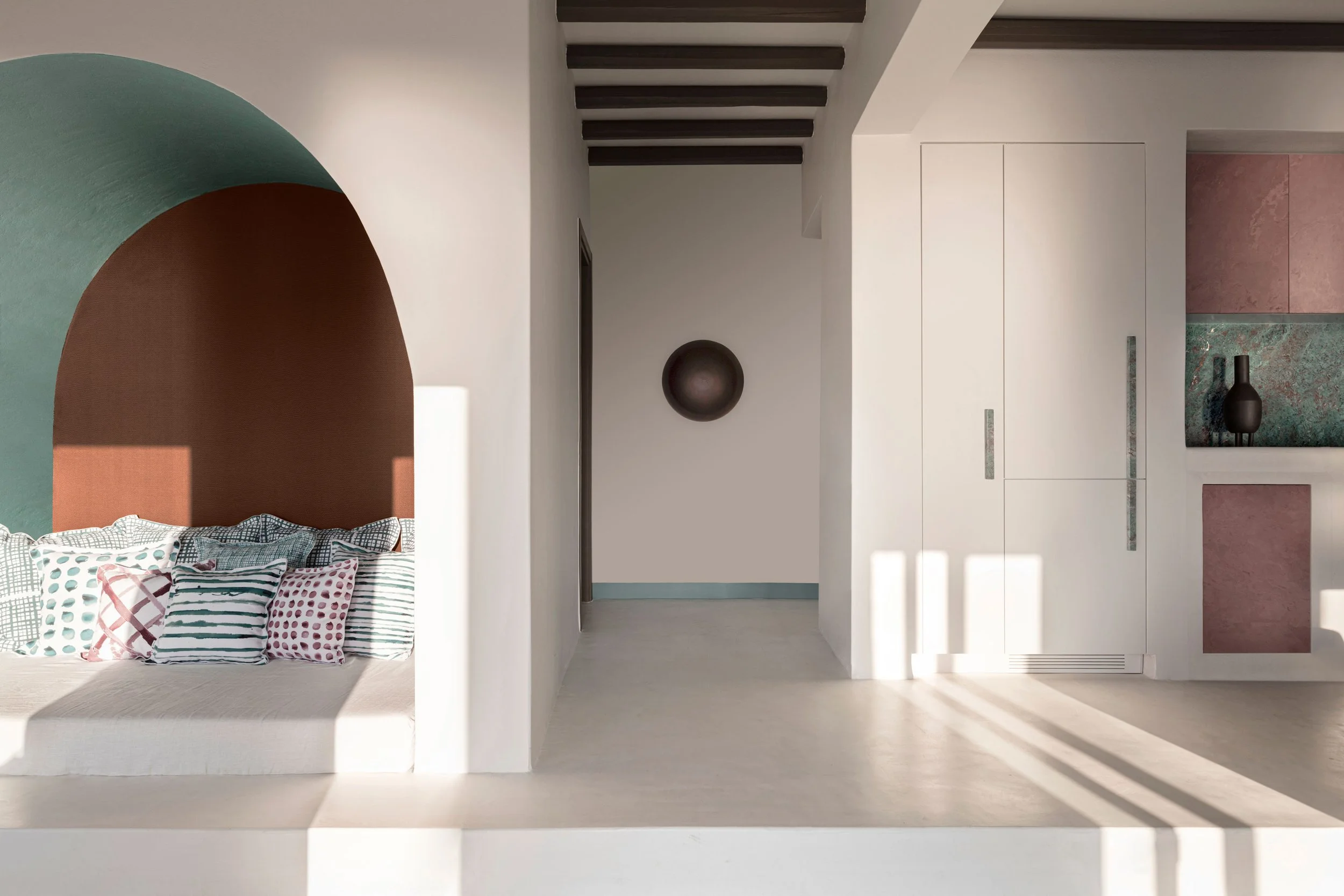 Minimalist interior with bed featuring patterned pillows, a white wall, a dark round decorative piece, white cabinetry, and pink and green tiled accents. Sunlight streams in creating shadows.