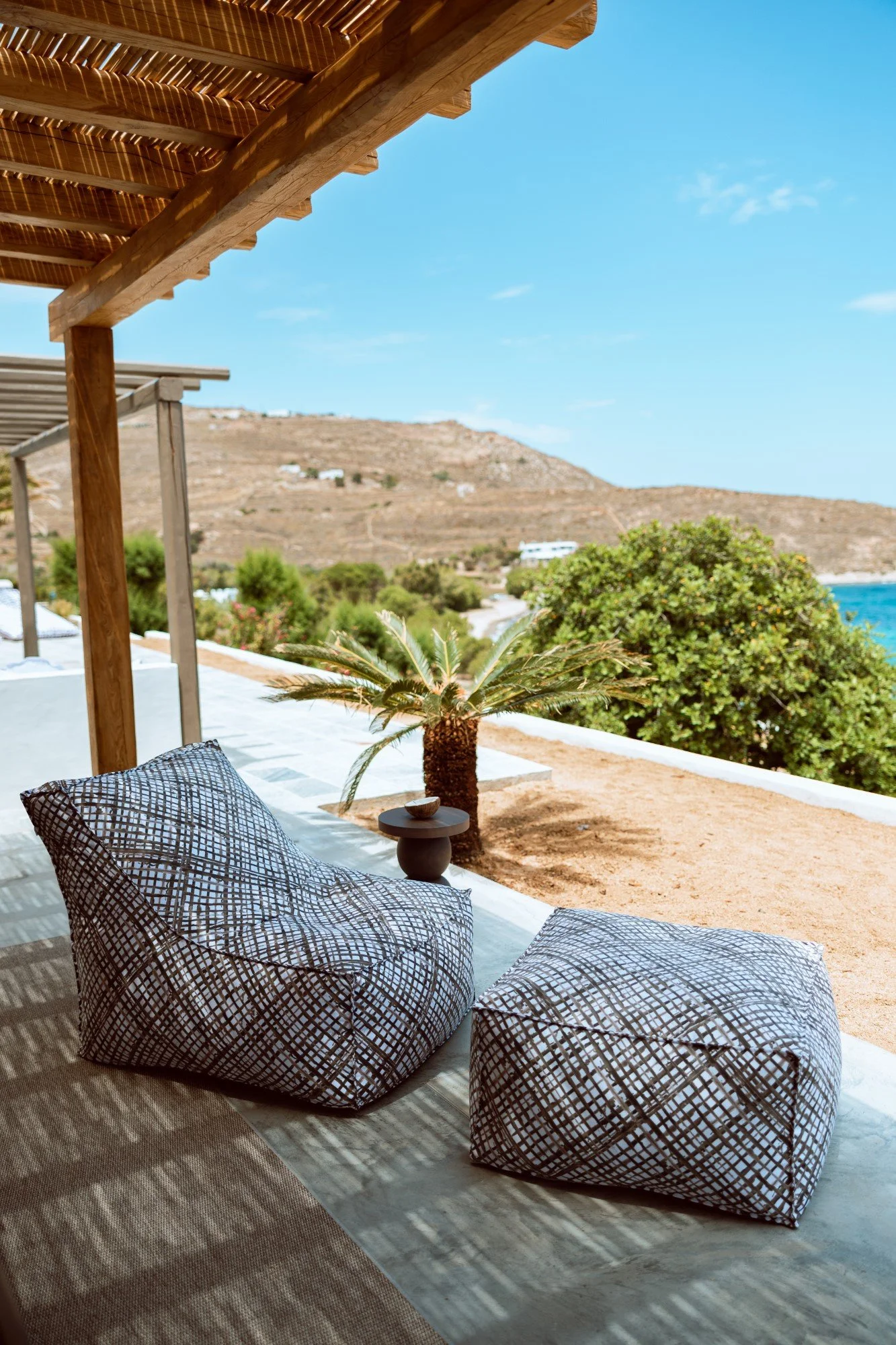 Outdoor seating area with patterned cushions, a small black table, a potted palm, and a scenic hillside view with bushes, clear blue sky, and part of a beach.