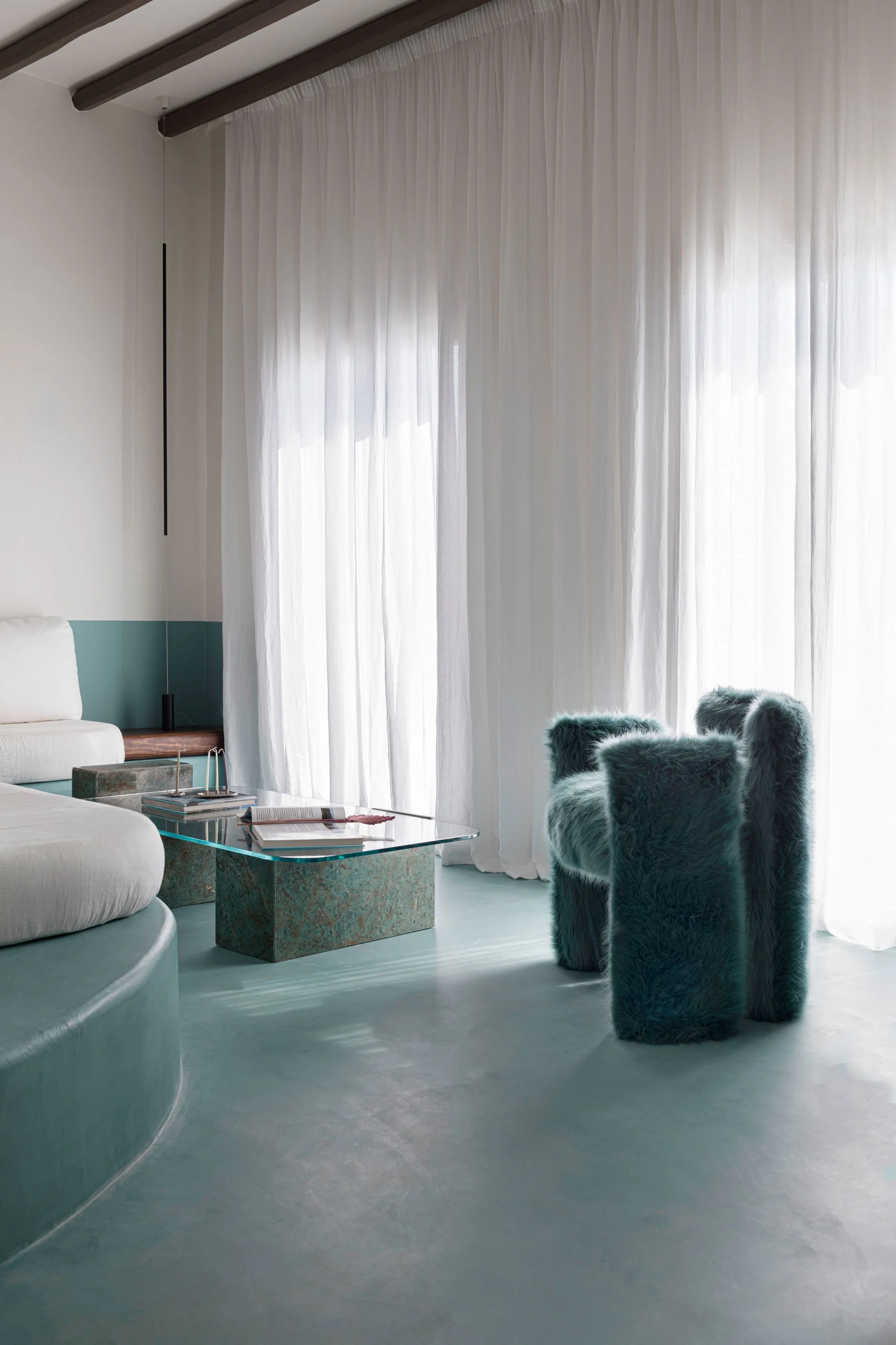 Living room with sheer white curtains, a white sofa, a glass-topped coffee table with books, and three plush, teal-colored furry chairs.