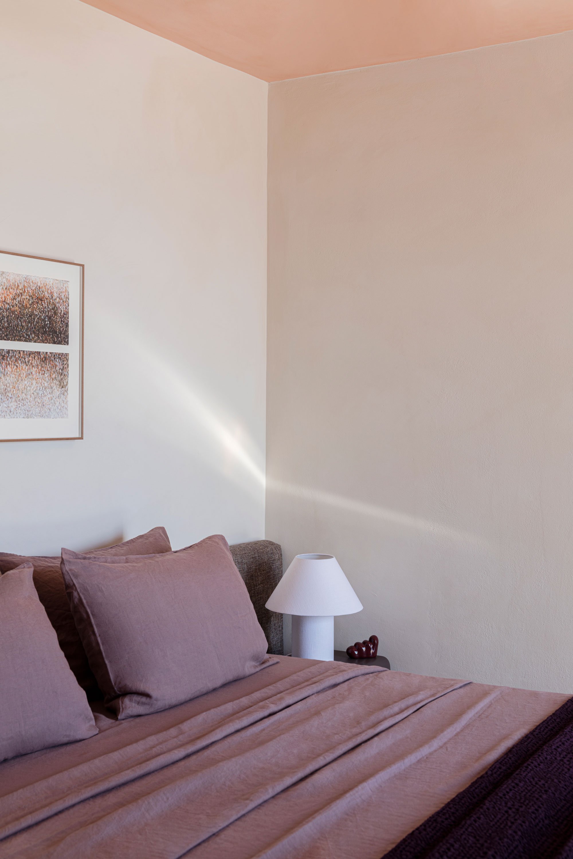 A bedroom corner with a bed having mauve pillows and a mauve blanket, a white bedside lamp, and a framed abstract artwork on the wall.