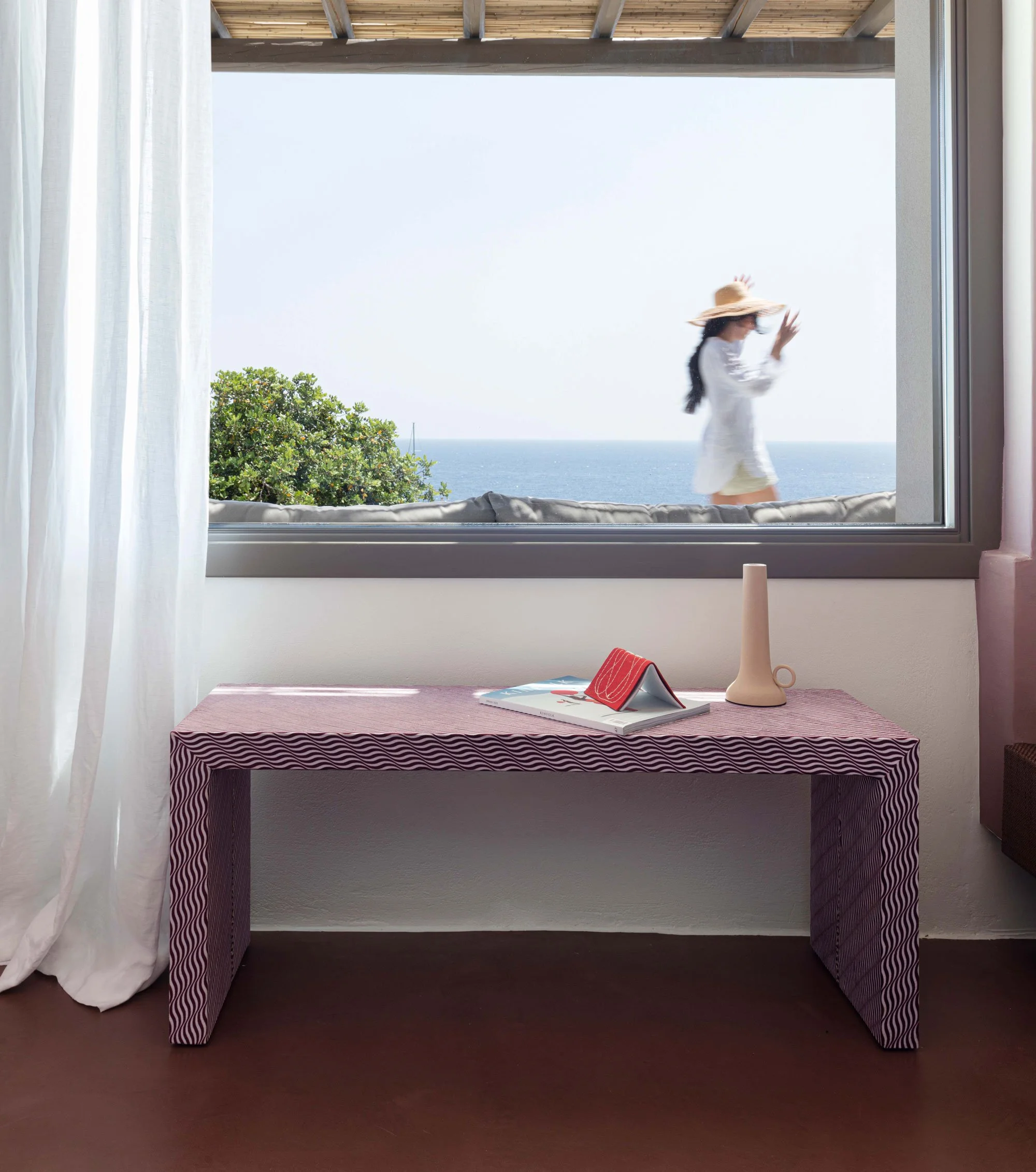 View through a window showing a woman in a white dress and wide-brimmed hat walking outside near the ocean, with trees and blue water in the background.