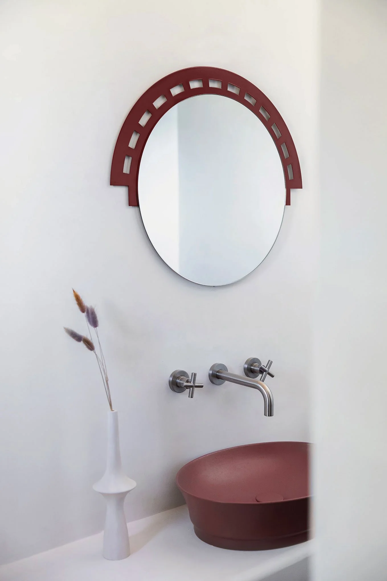 A bathroom with a round mirror above a sink. The mirror has a red frame with rectangular cutouts. There are two silver wall-mounted faucets. A white vase with dried lavender flowers is on the counter.