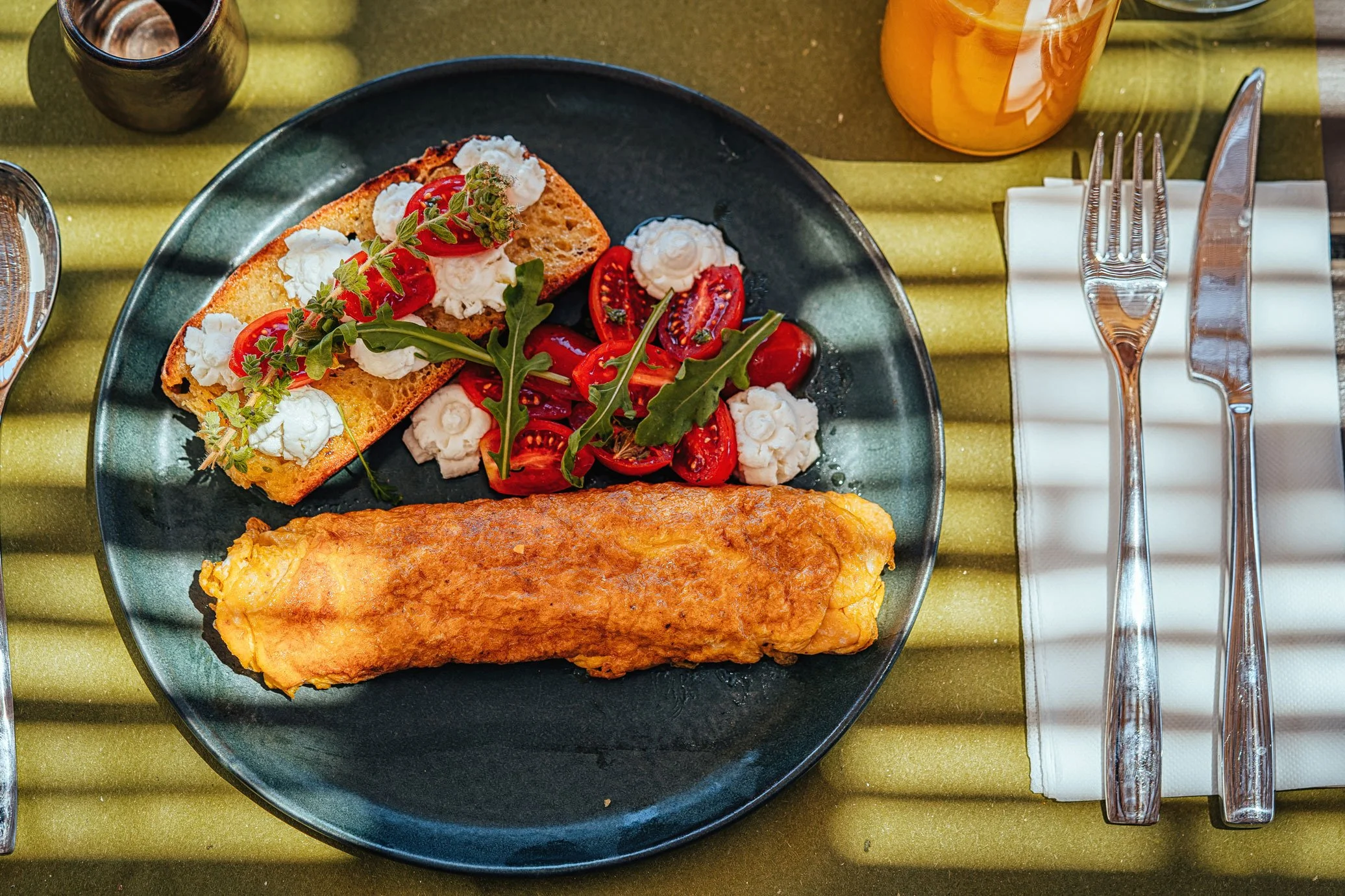 Plate with an omelette, toasted bread with cream cheese and cherry tomatoes, and a side salad with arugula and cherry tomatoes, on a green placemat.