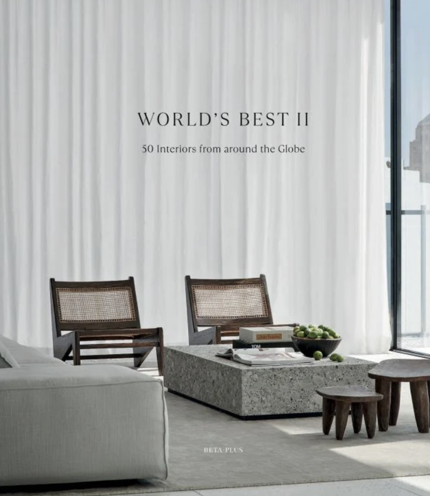 Cover of a magazine or book titled 'World's Best II' featuring 50 interiors from around the globe. The image shows a modern living room with white curtains, a white sofa, two wooden chairs with woven seats, a gray stone coffee table with books and a bowl of green apples, and small wooden stools. A sliding glass door on the right reveals cityscape views.