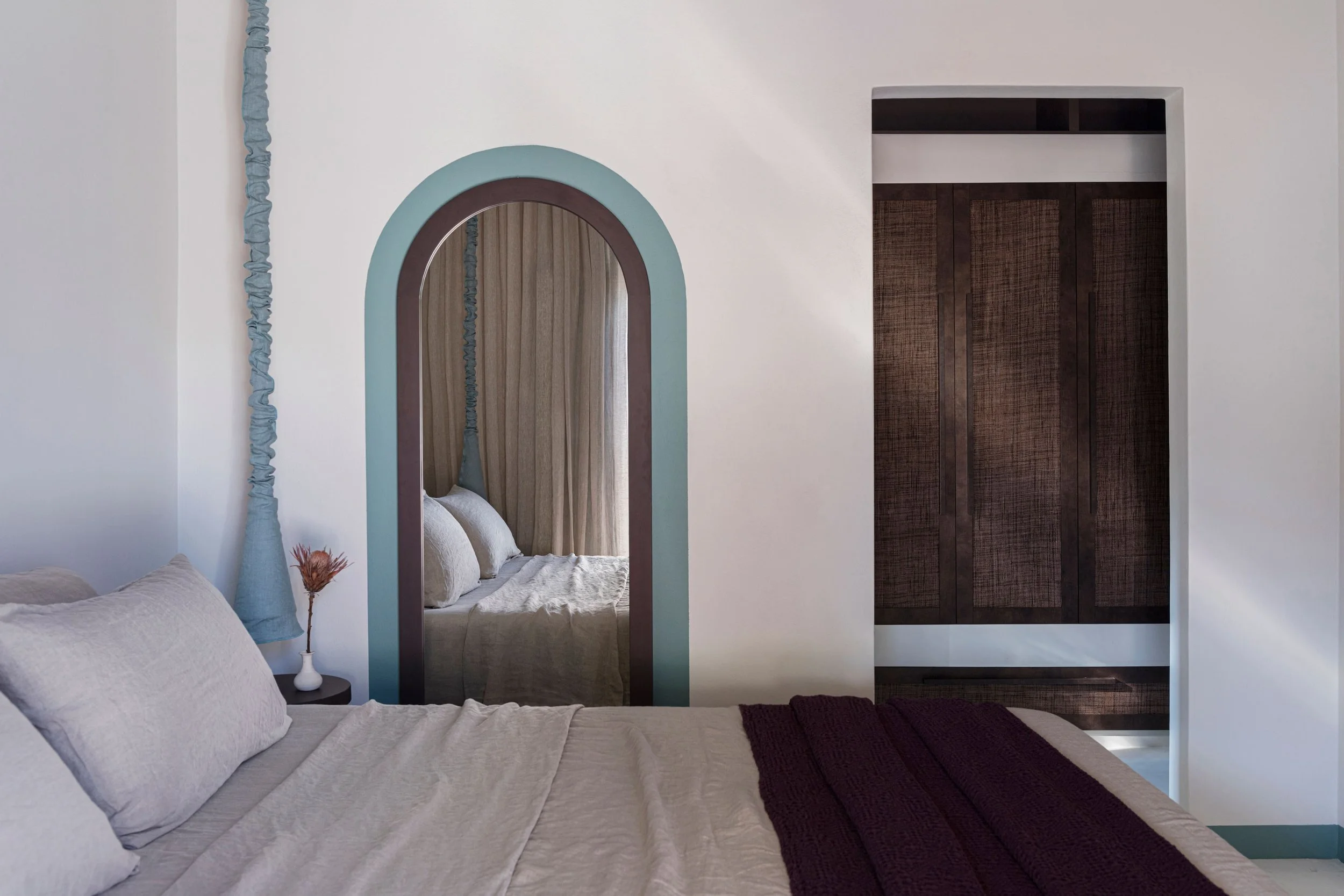 A bedroom with a bed, pillows, a bedside table with a vase and flower, a mirror, and a wardrobe.