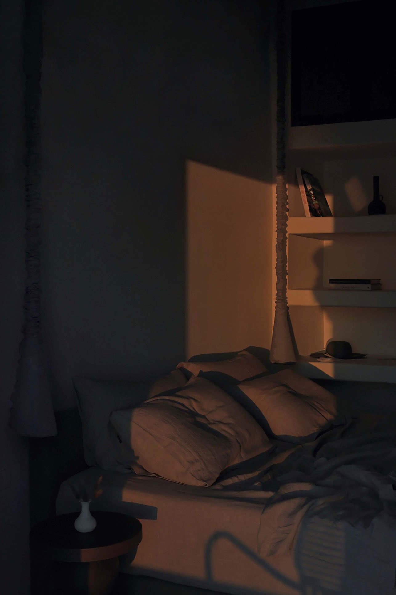 A cozy bedroom scene with a bed, pillows, and a nightstand featuring a small vase. The wall behind the bed has a harsh shadow cast by a window blind, with parts of a bookshelf visible on the right side of the image, illuminated by warm sunlight.