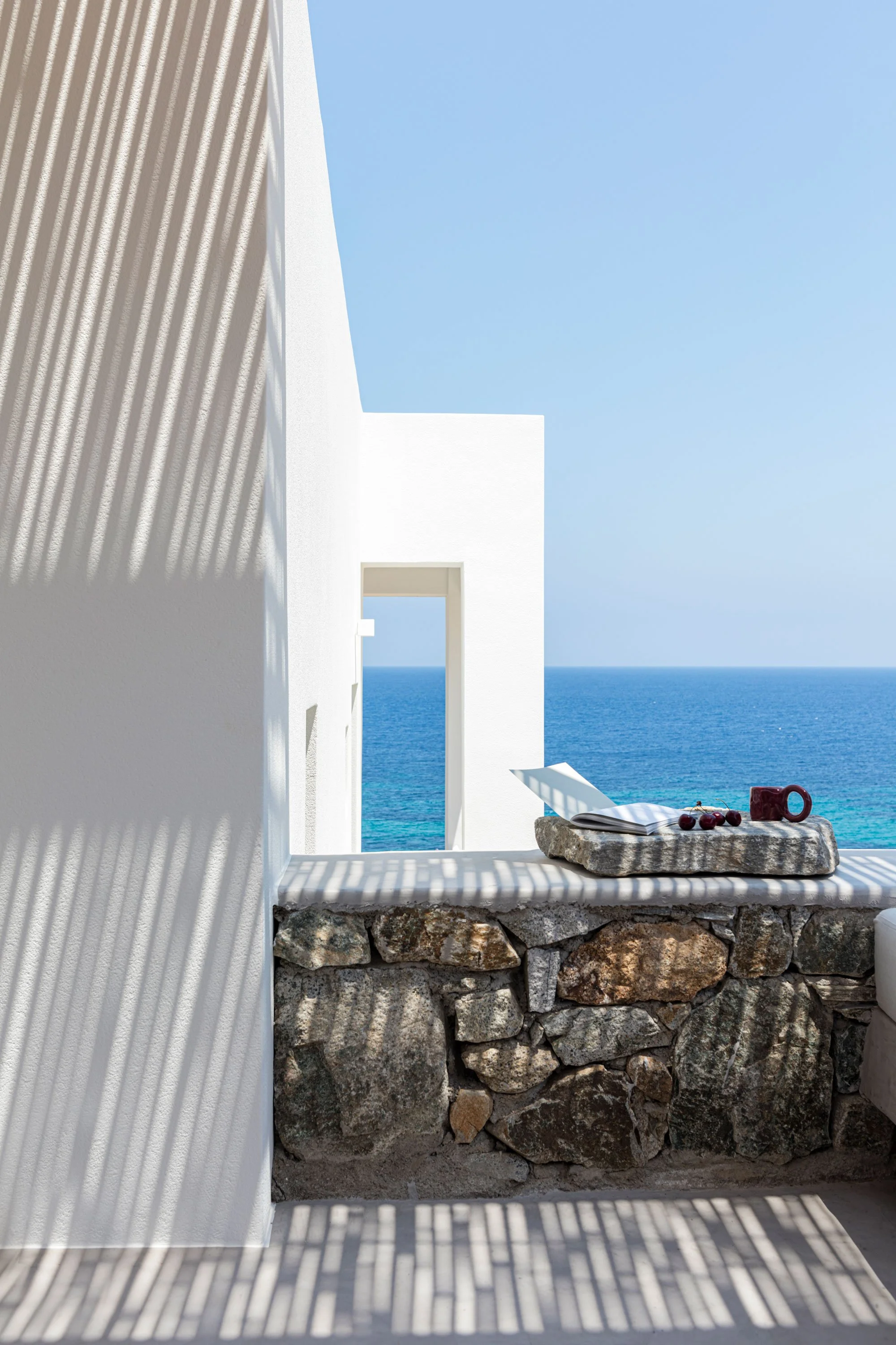 A white modern building overlooking the ocean with shadows cast by a pergola, a stone ledge with an open book, cherries, and a coffee mug on it.