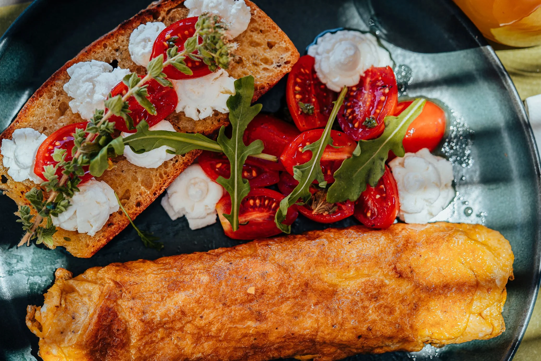 Omelette, tomato salad with fresh herbs and arugula, and toasted bread topped with cheese and herbs on a black plate