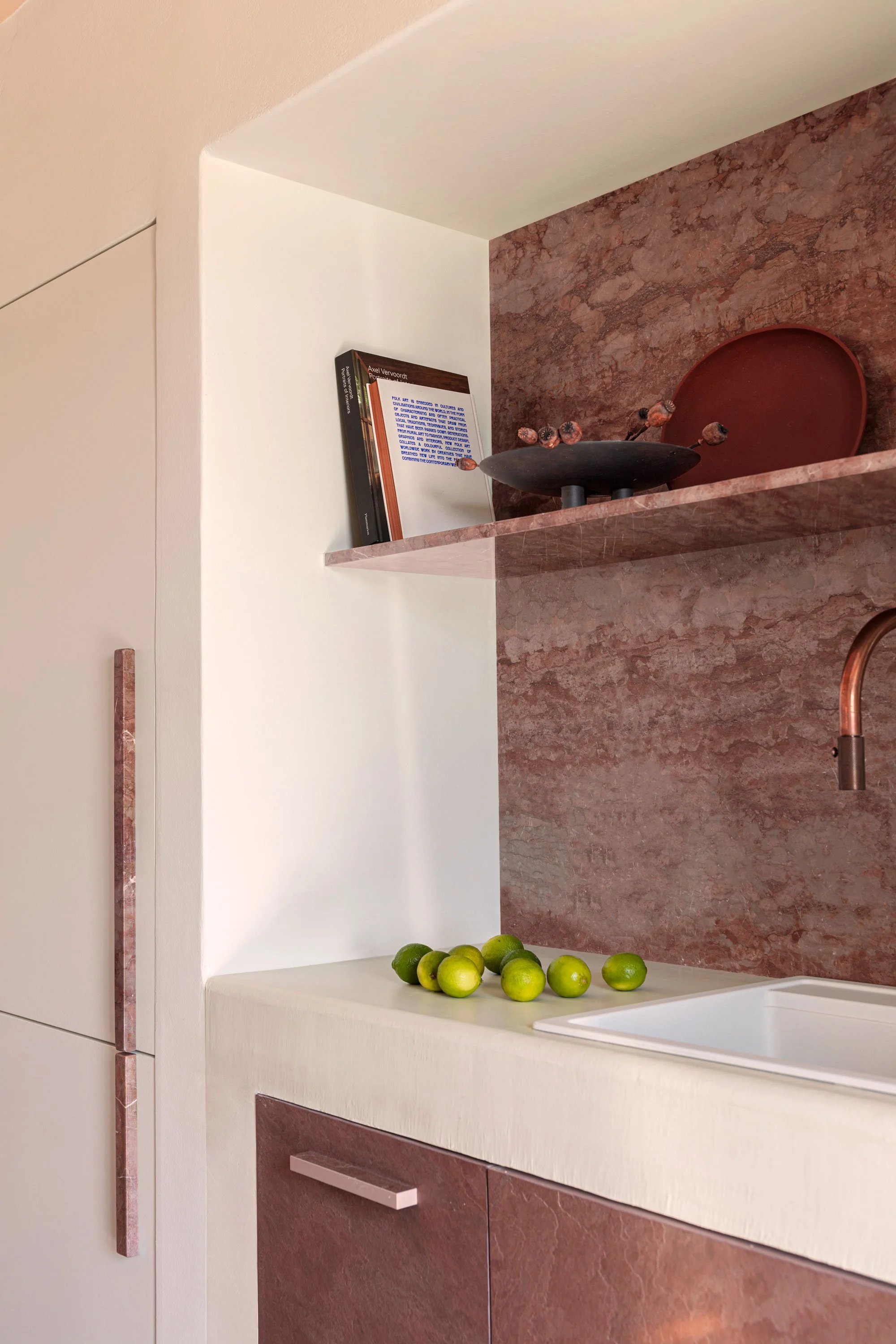 Corner of a modern kitchen with a marble countertop, a sink, and a series of limes on the counter. There are floating shelves with decorative objects and a book.