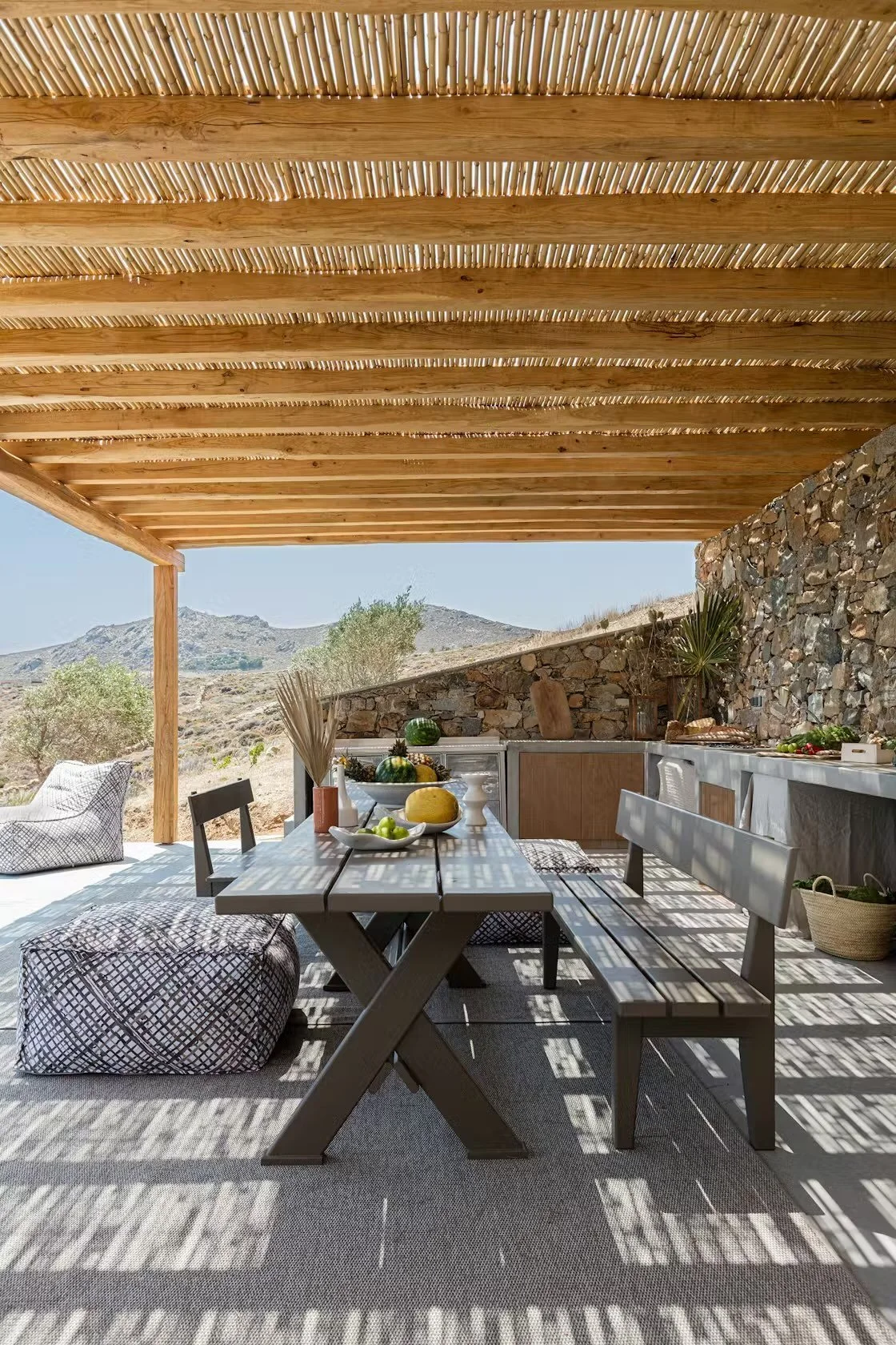 Outdoor dining area with wooden table, benches, and chairs under a slatted wooden roof, overlooking a desert landscape with hills and sparse vegetation.