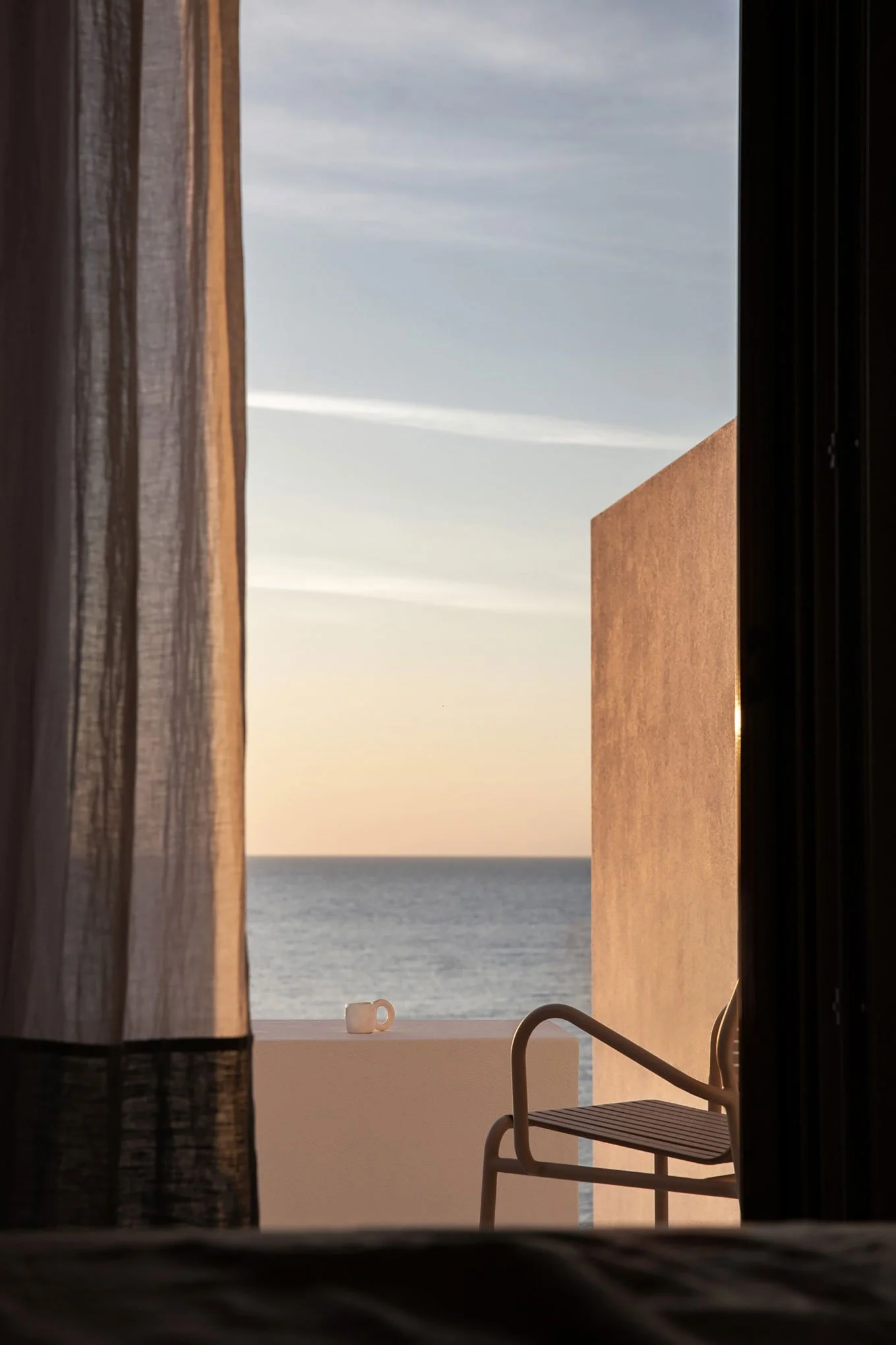 View of the ocean through a partially open window with sheer curtains, a patio chair, and a coffee mug on the outside railing at sunset.