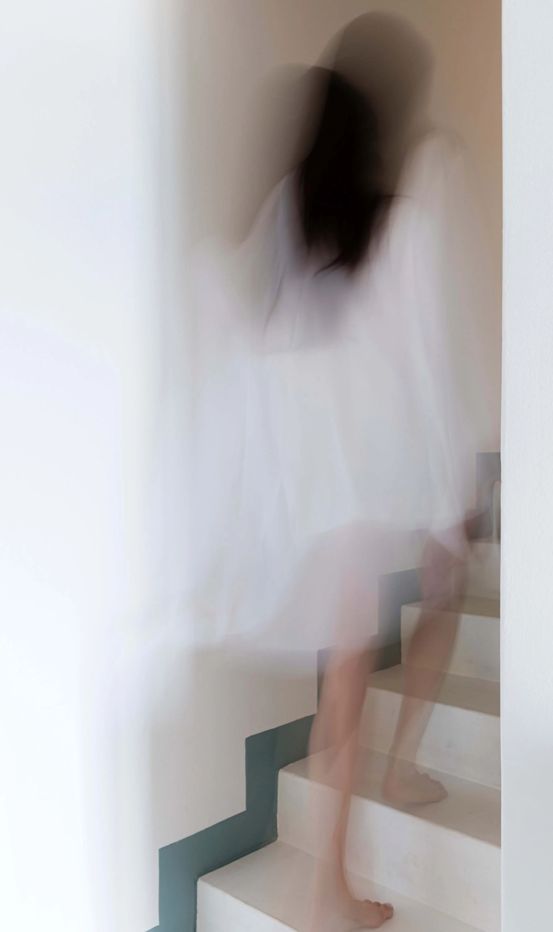 Blurry photo of a woman walking up stairs, wearing a white dress, with dark hair, in a minimalistic space with white walls.