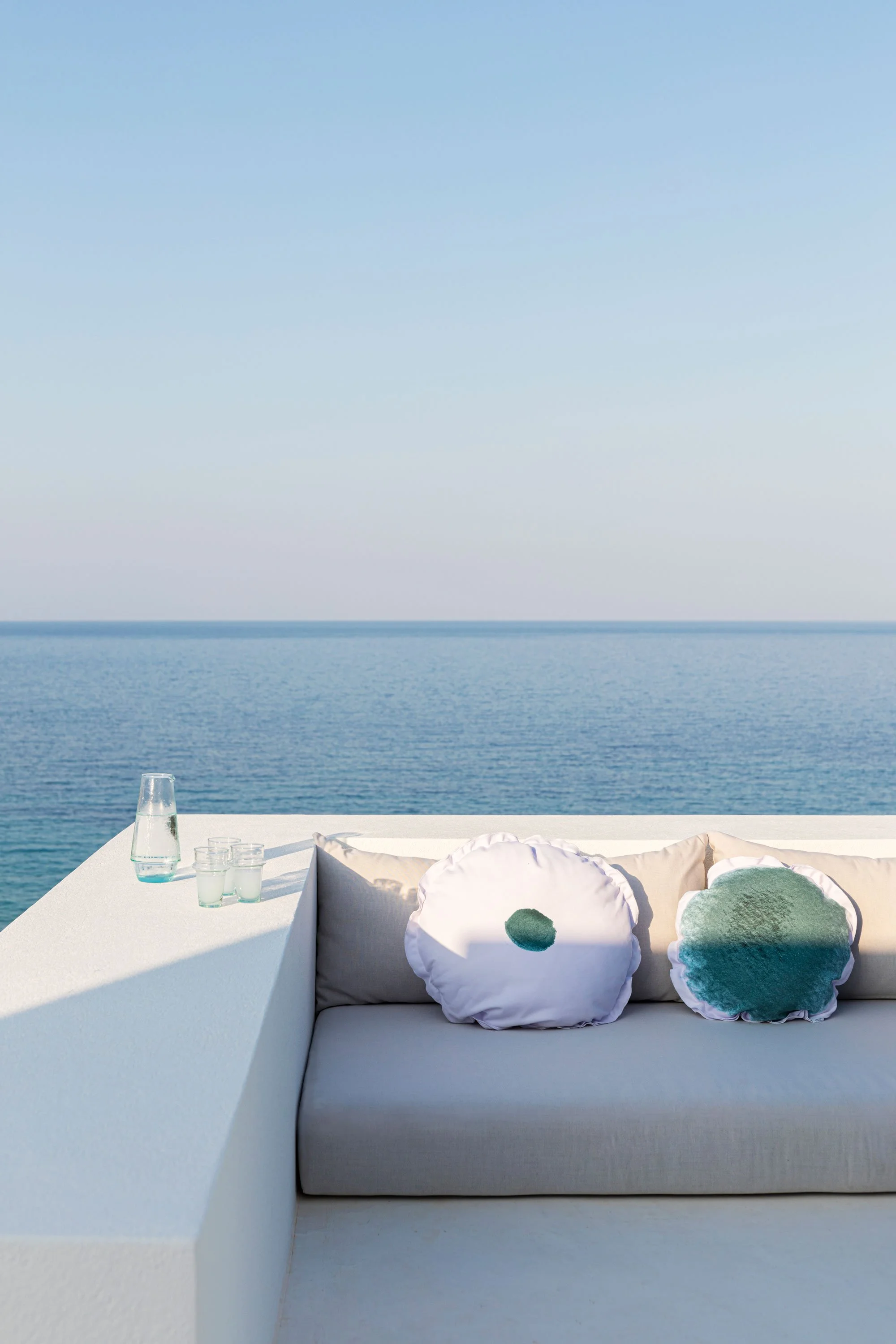 Outdoor seating area overlooking the ocean with decorative pillows and a table with drinks and a water pitcher.