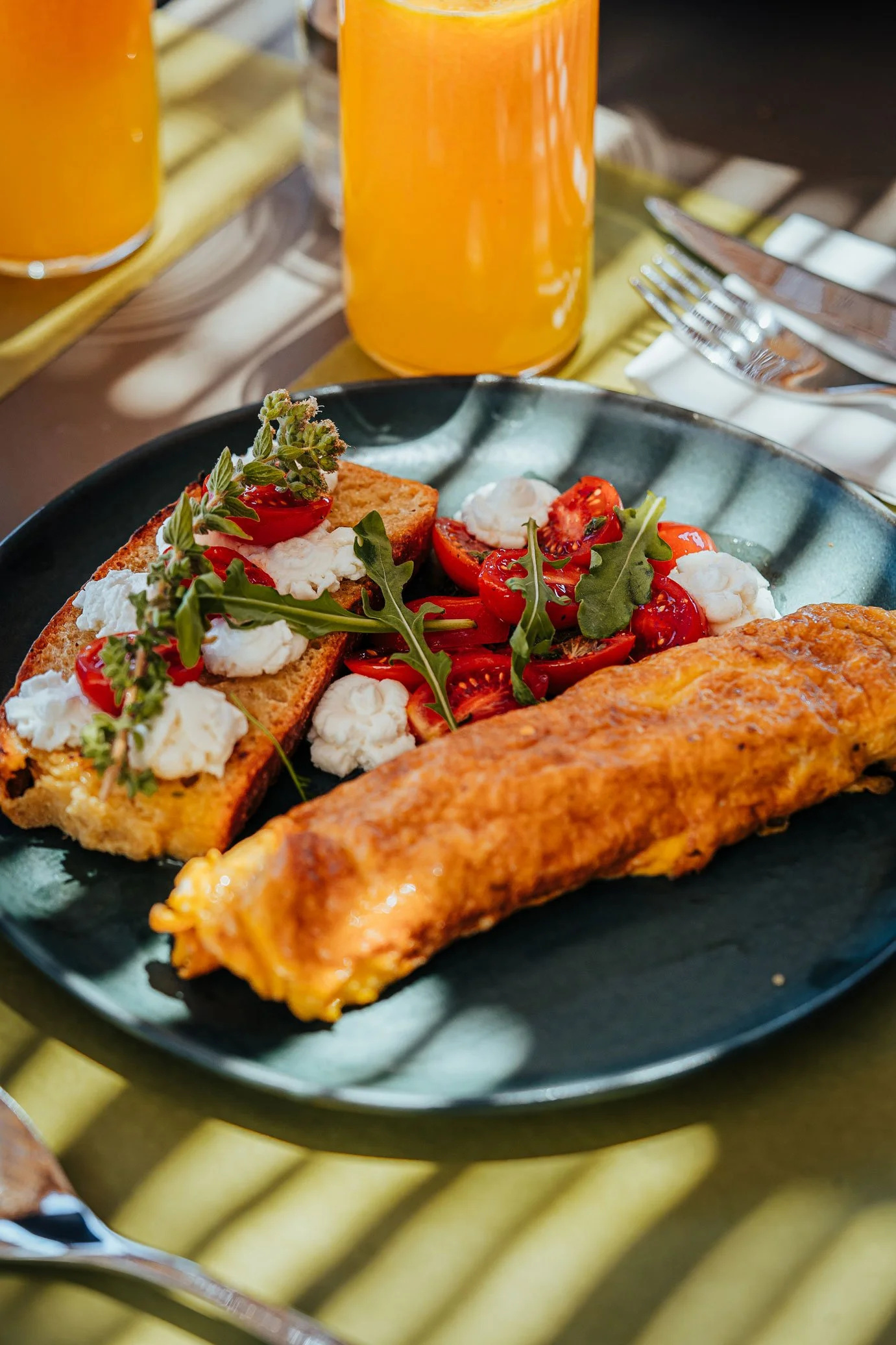 A plate of breakfast featuring a scramble omelet, a tomato and arugula salad, and a slice of bread with cheese, herbs, and cherry tomatoes, with a glass of orange juice in the background.
