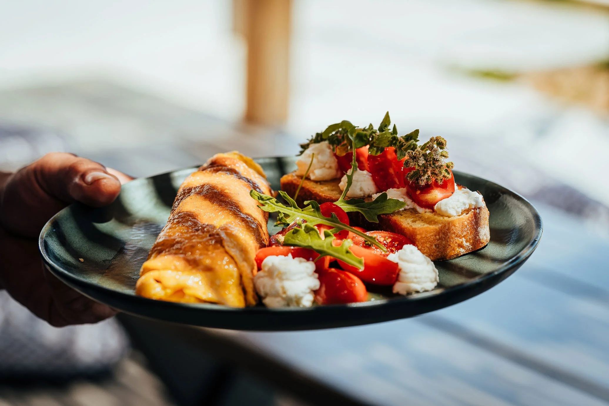A person holding a black plate with a slice of omelette, bruschetta topped with tomatoes, cheese, and greens, and some fresh arugula and cauliflower.