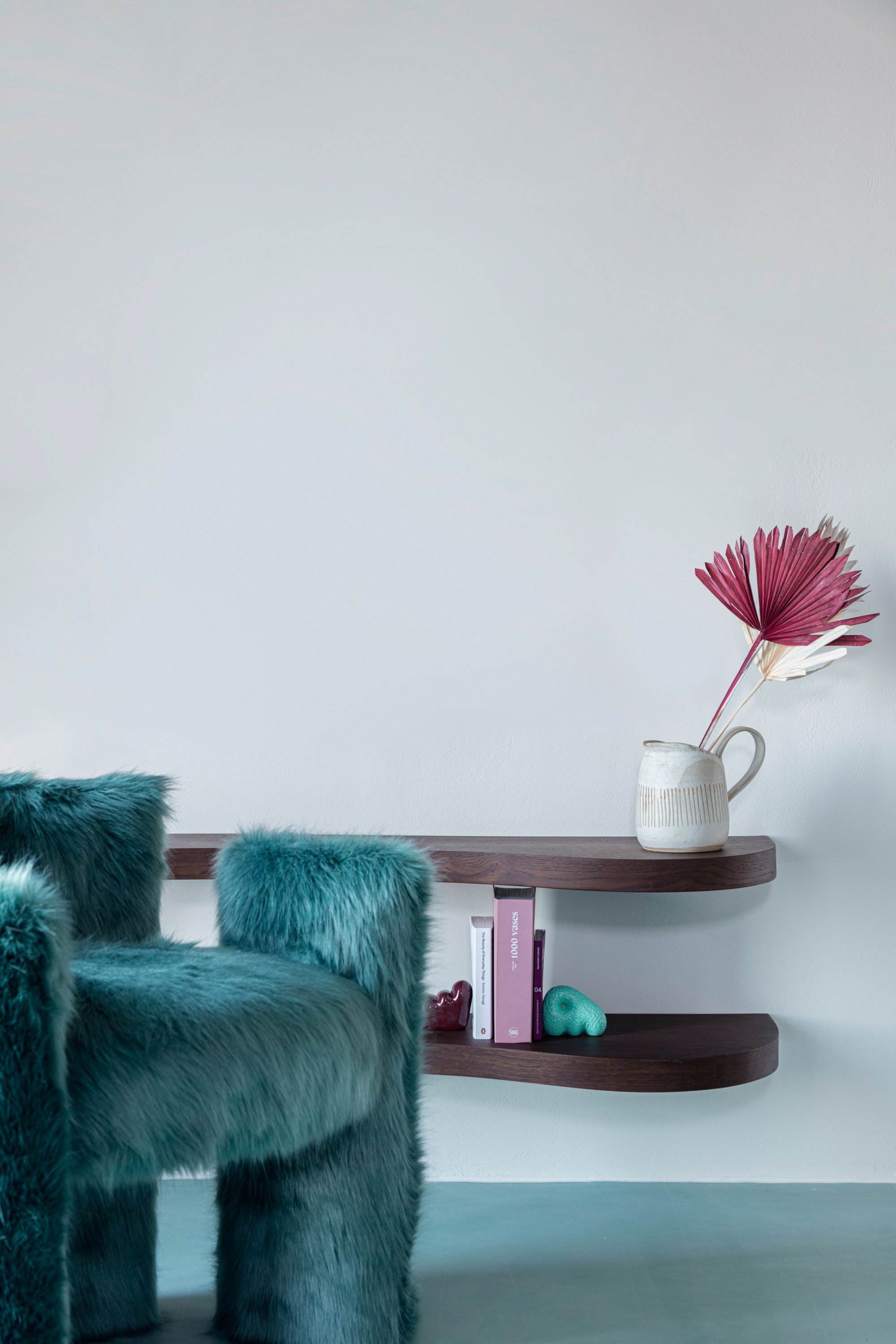 A teal furry armchair with matching furry pillows next to a dark wooden floating shelf holding a white vase with pink and white dried leaves and some books and decorative objects underneath.