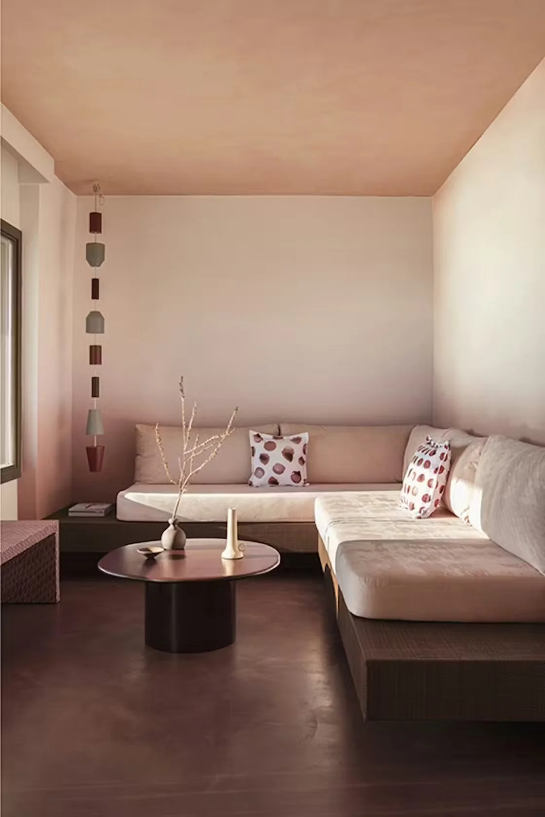 Minimalist living room with a beige L-shaped sofa with patterned cushions, a dark round coffee table with decorative vases, a textured square ottoman, and hanging pendant lights near a large window.