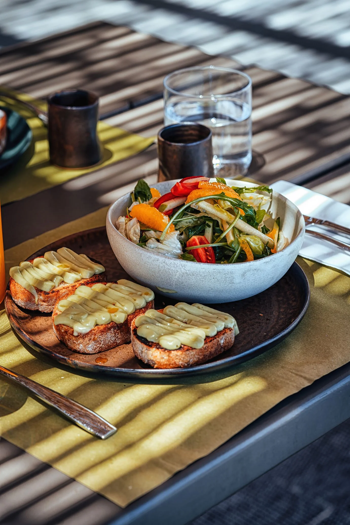 A plate with four pieces of toasted bread topped with cheese, a bowl of fresh mixed salad with tomatoes, cucumbers, and orange slices, a glass of water, and two dark cups on a table outdoors.