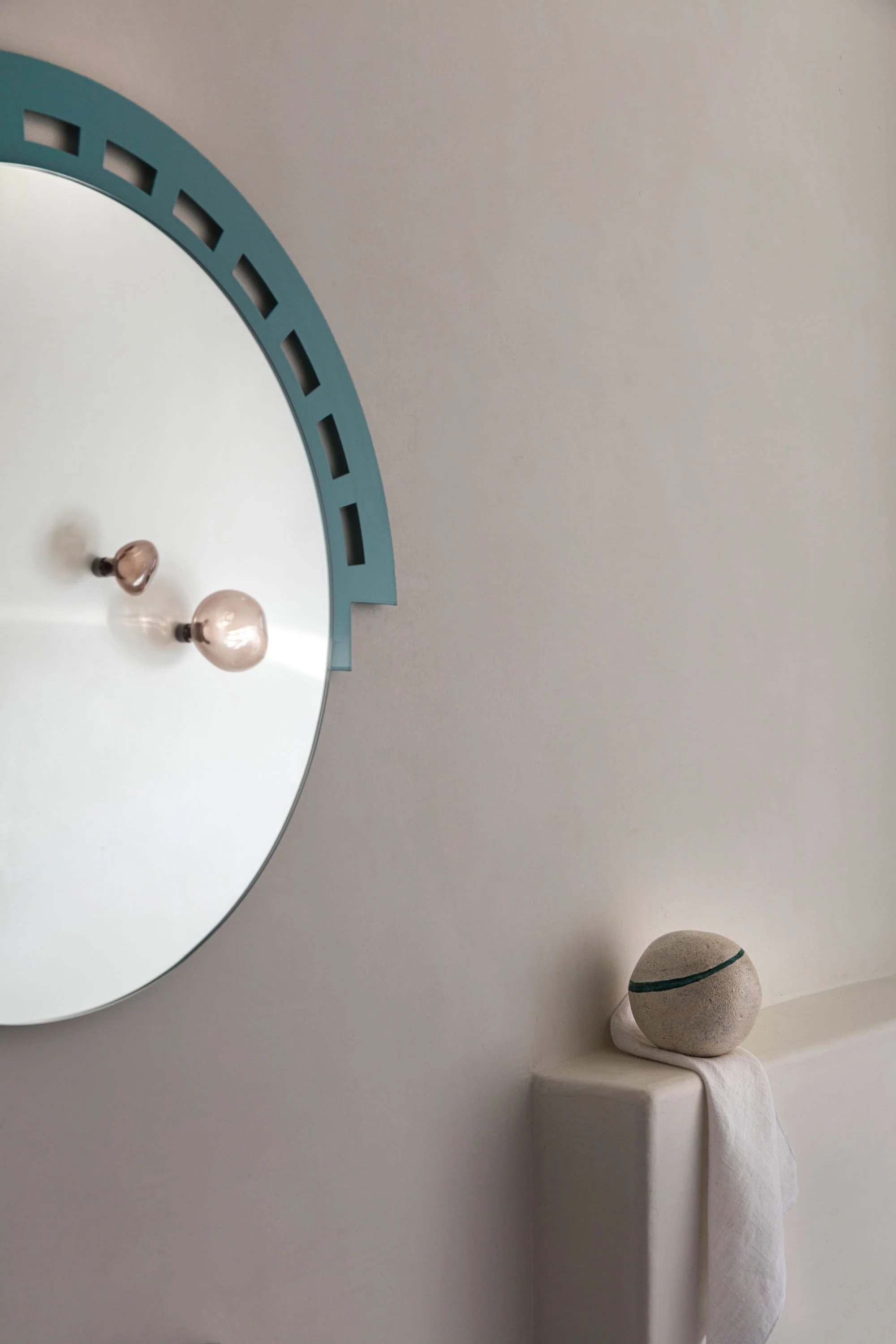 Decorative interior wall with a round mirror featuring a blue frame, a ceiling light with two bulbs, a white cloth, and a round stone with a green stripe resting on a ledge.
