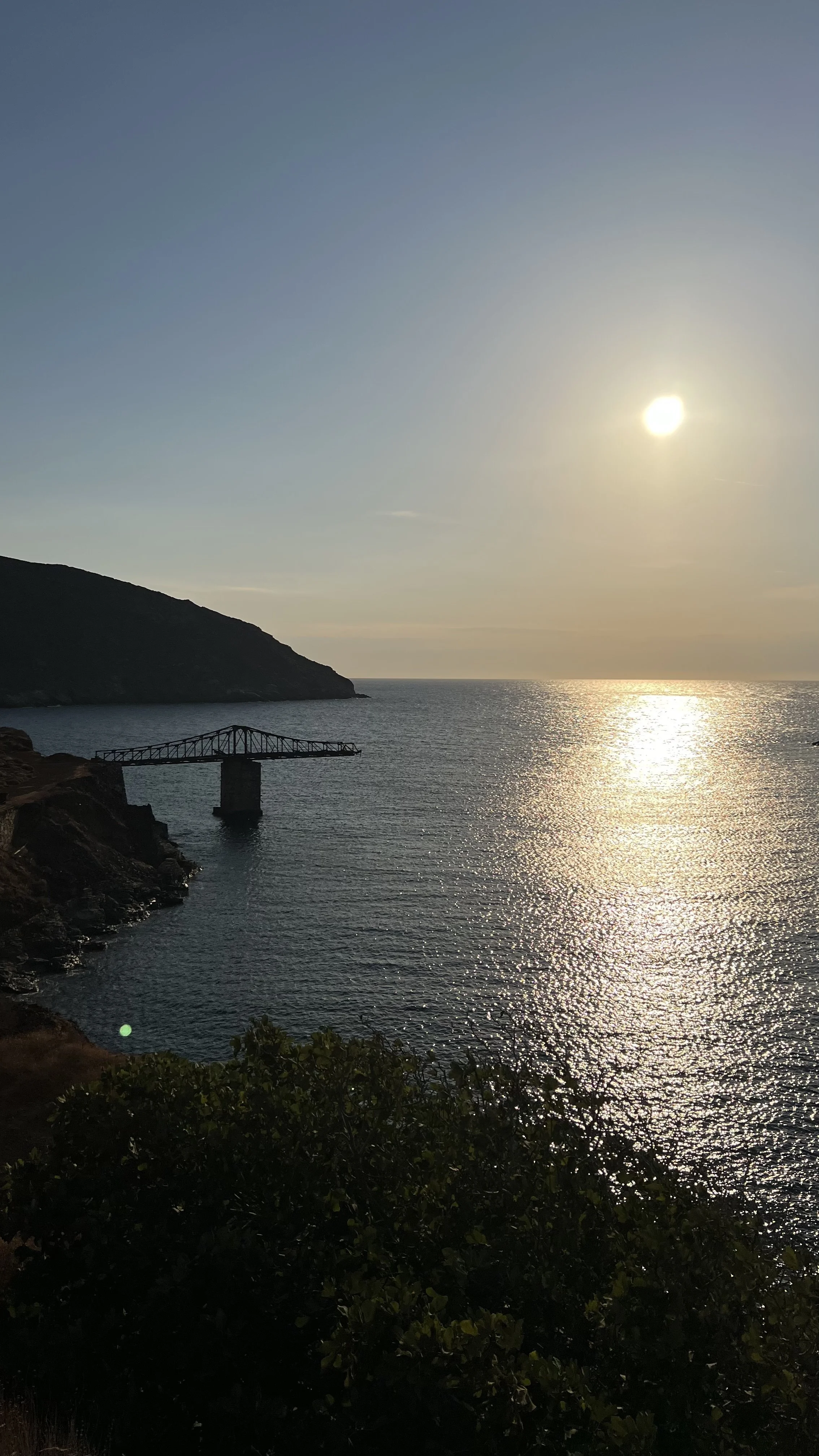 Sunset over the ocean with a rocky shoreline, a small pier structure, and a hill in the background.
