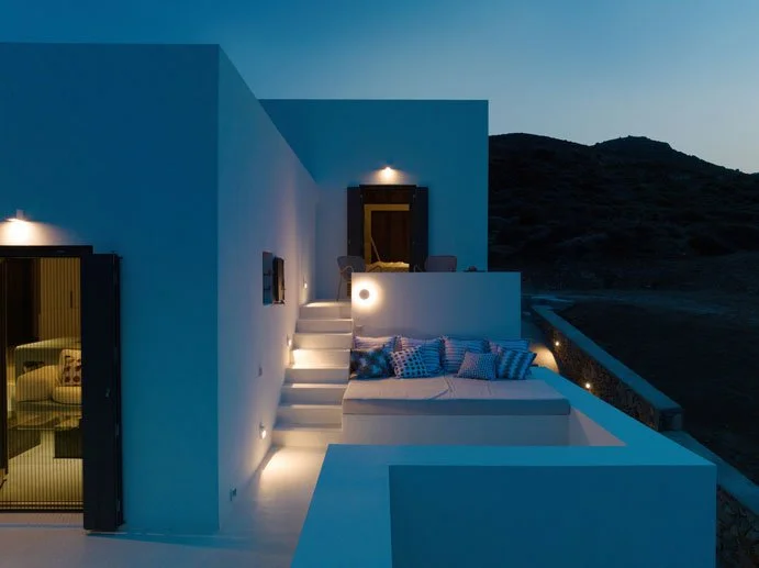 Nighttime view of a modern white building with outdoor stairs, a balcony with chairs, and a lounge area with pillows, illuminated by warm lights, overlooking a dark landscape.