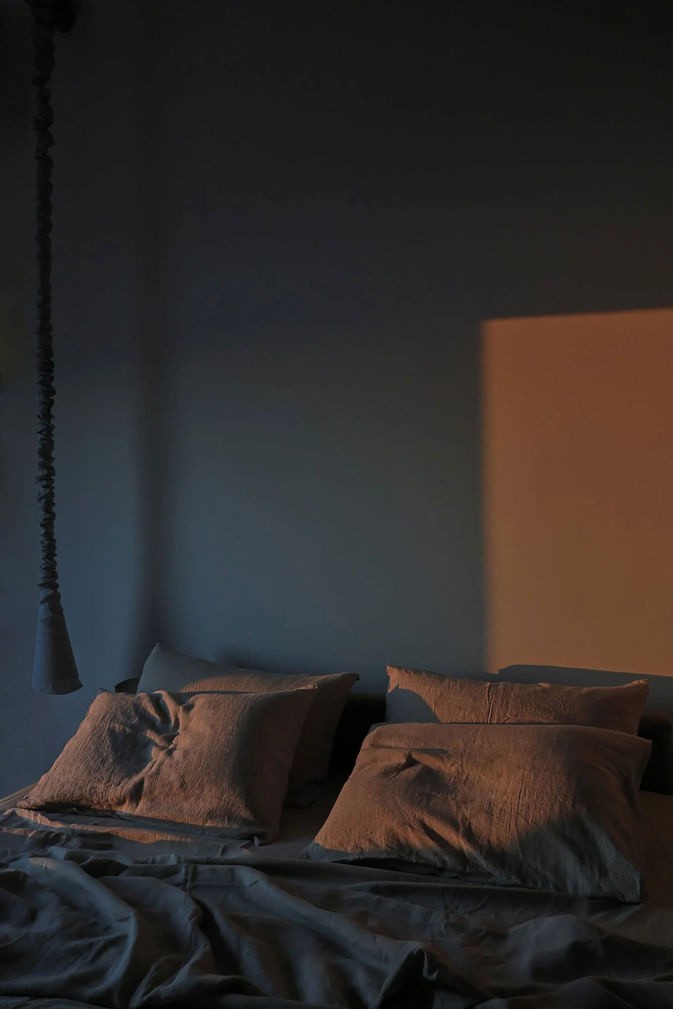 A dimly lit bedroom with a bed and rumpled pillows, illuminated by warm sunlight casting shadows on the wall.