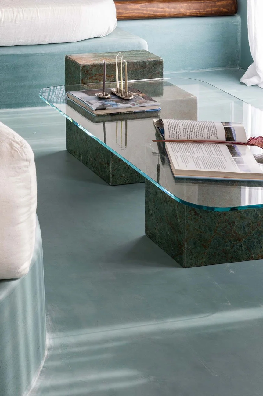 A glass coffee table with stone legs in a bright living room, holding magazines, an open book, and a candle holder with three unlit candles.