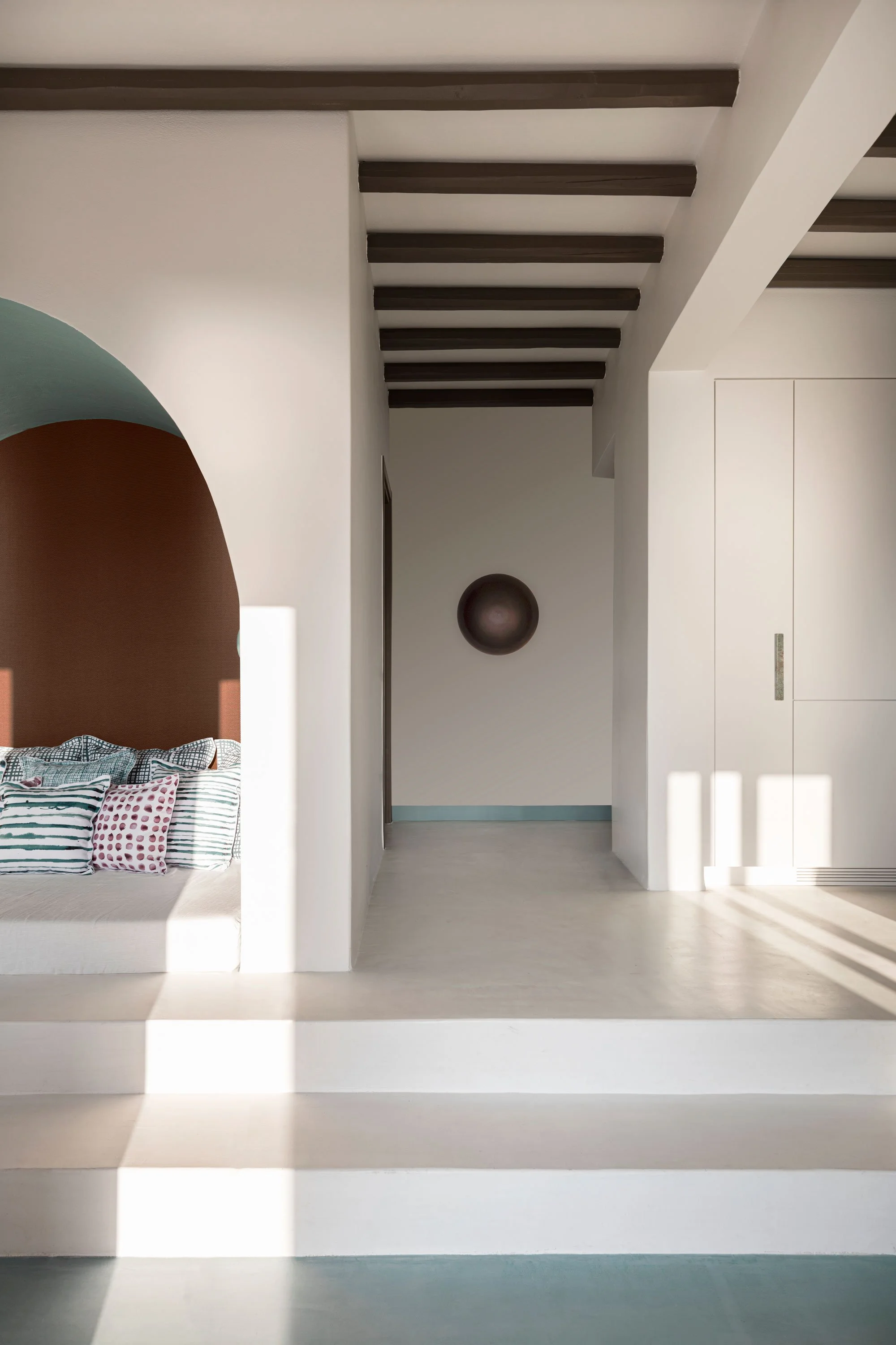 Minimalist interior scene with a white wall, a dark circular sculpture, and wooden ceiling beams. Part of a bedroom with a bed and colorful pillows is visible on the left.