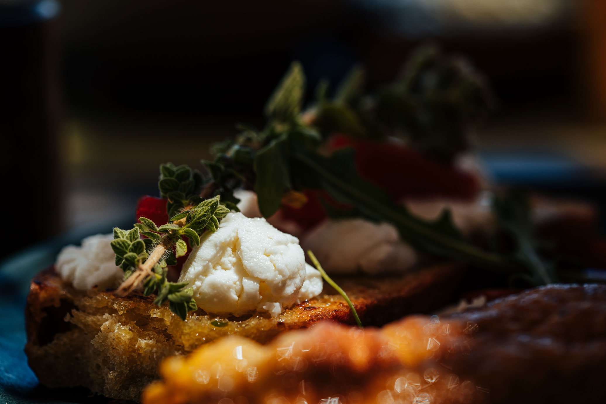 Close-up of a slice of bread topped with cheese, herbs, and garnished with fresh thyme, with other food items blurred in the background.