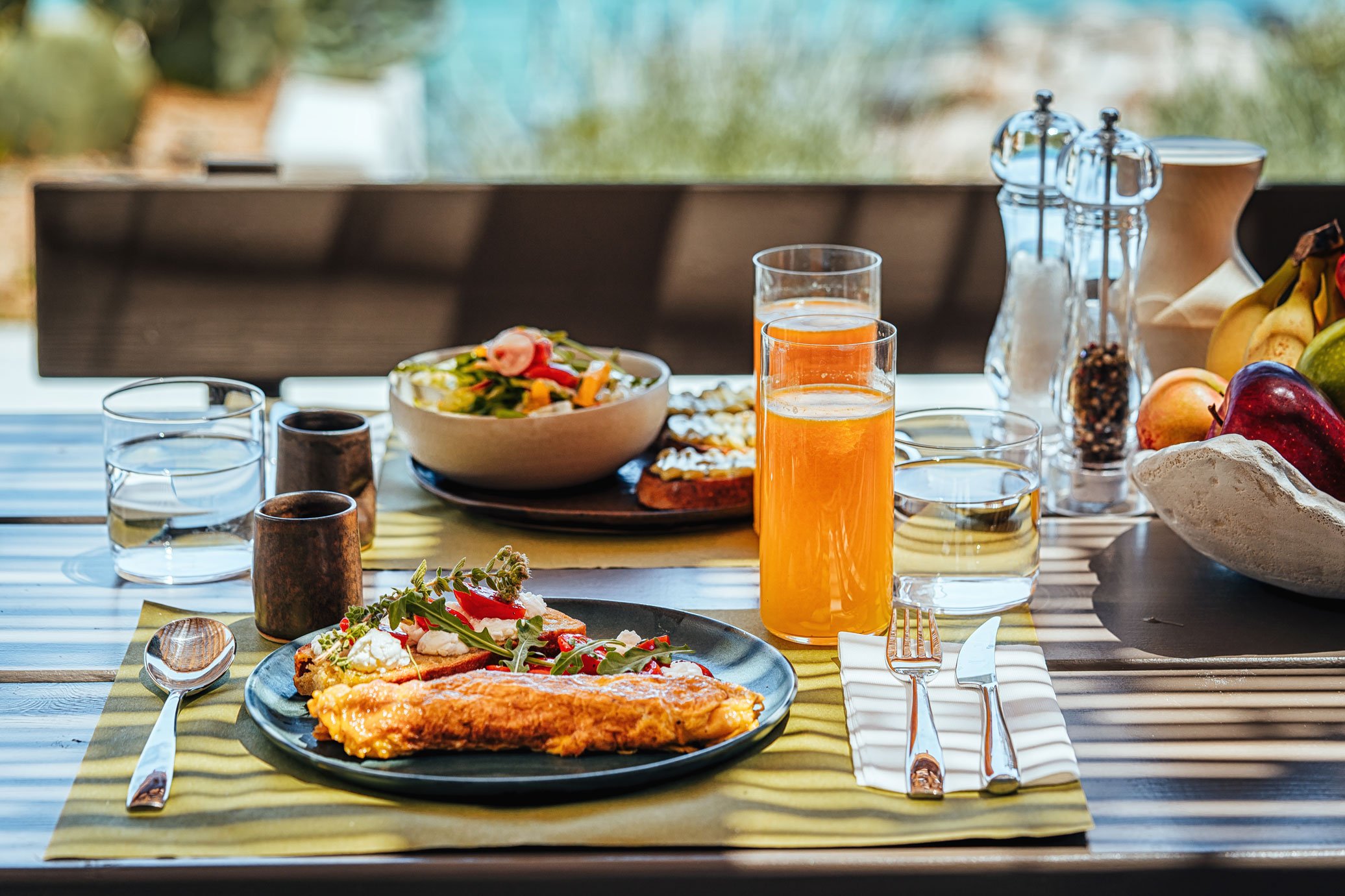 Table set with breakfast including a plate with fried fish, salad, glasses of orange juice, water, a bowl of salad, a bowl of fruit, salt and pepper shakers, and cutlery on striped cloth placemats.