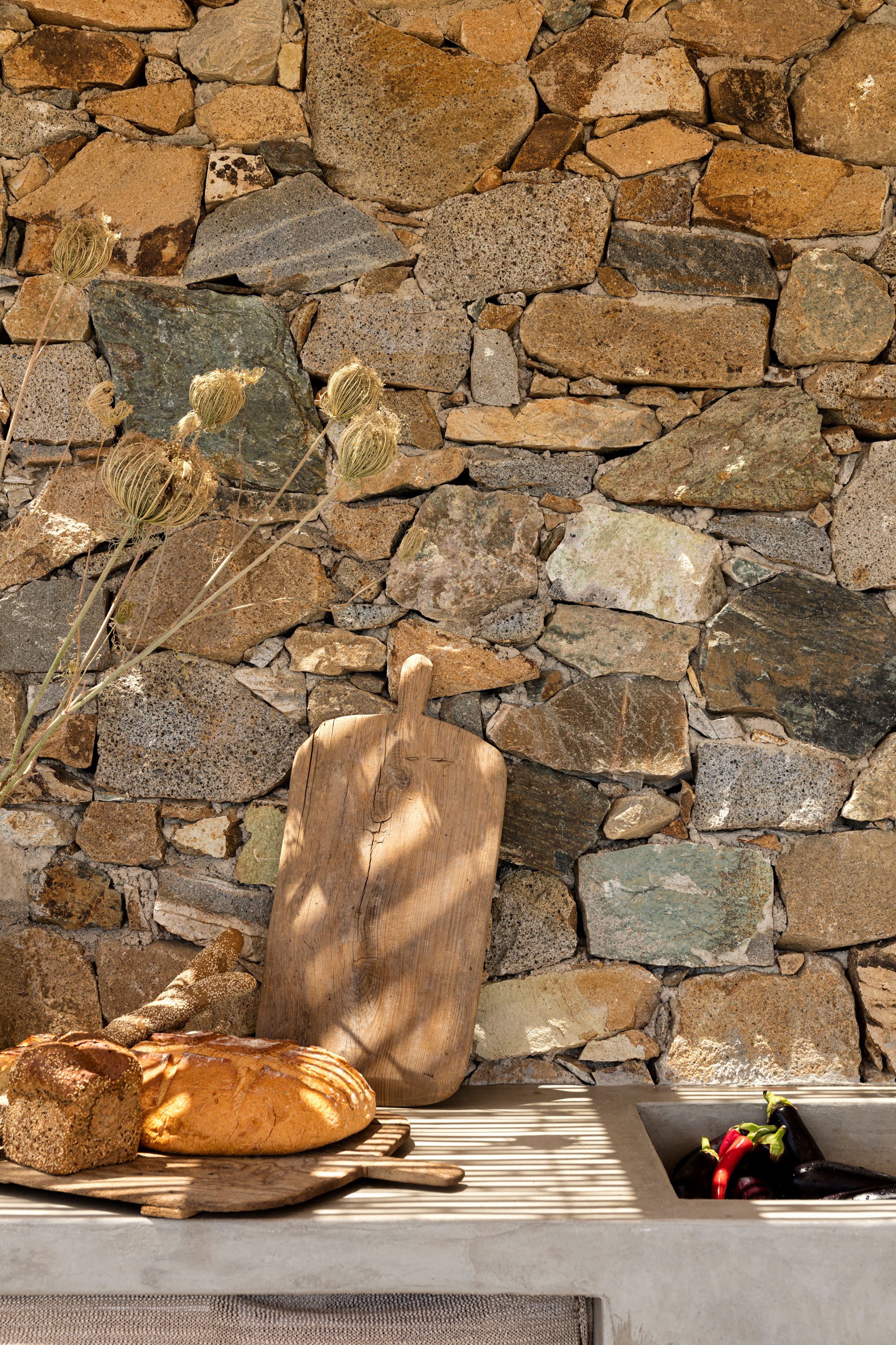 A rustic kitchen countertop with a stone wall background. There is a wooden cutting board, a loaf of bread, slices of bread, and a bunch of dried flowers on the counter. A sink with dark purple and red chili peppers is also visible.