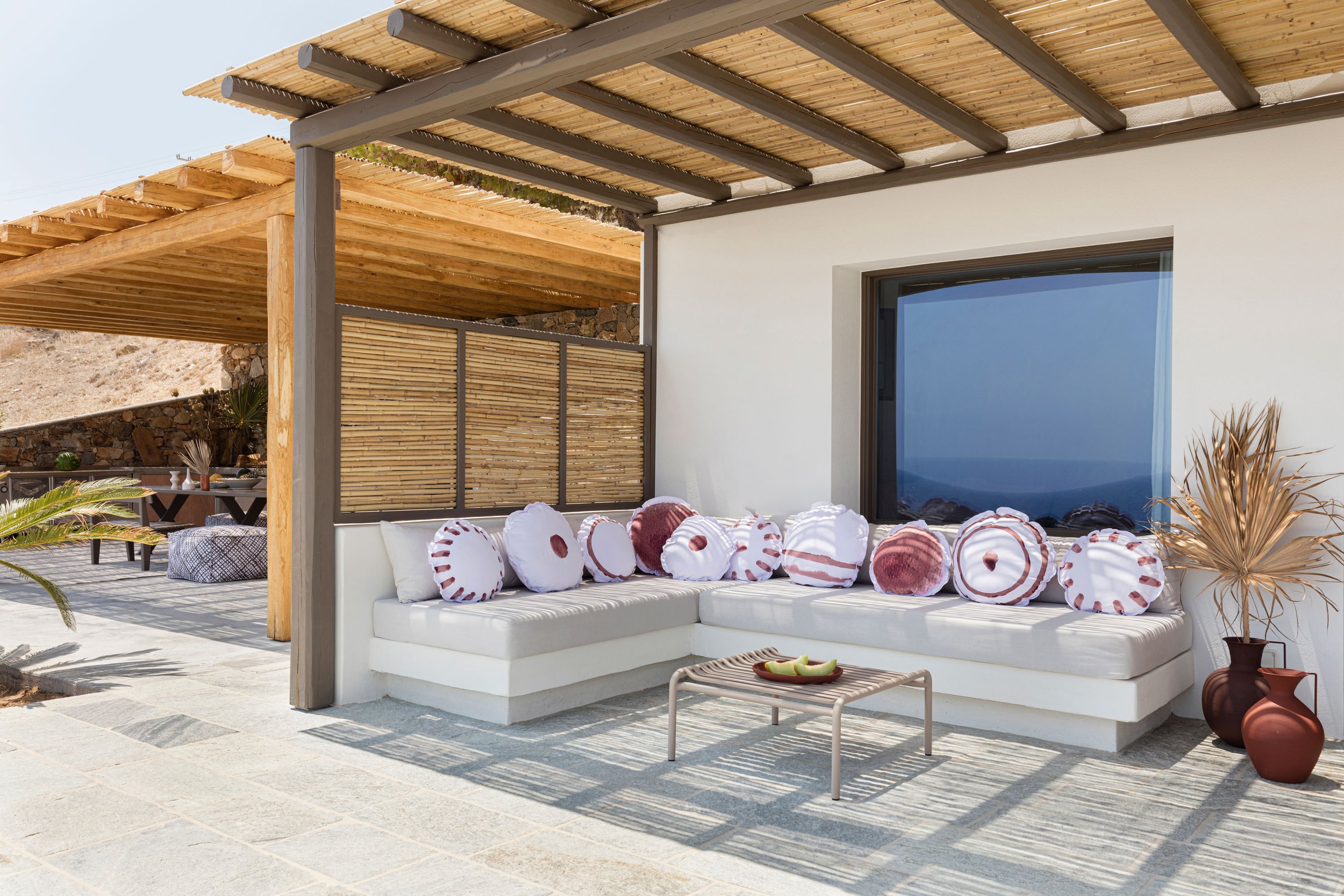 Outdoor patio with white U-shaped cushioned seating adorned with decorative pillows, a small coffee table with pineapple slices, and potted plants. The patio features a wooden pergola with slats and a white wall with a large window.