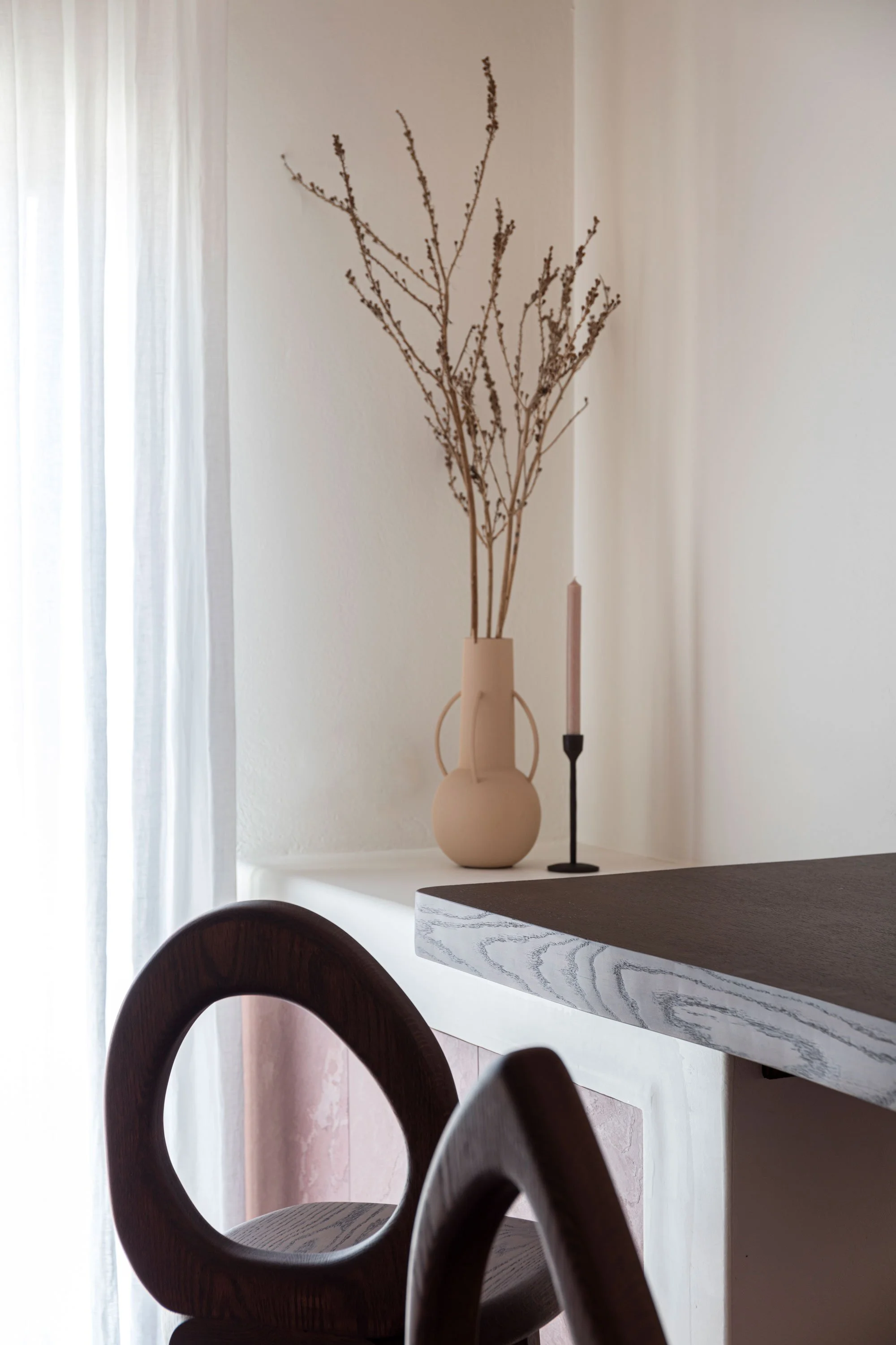 Interior scene with a white wall, a tall beige vase with dried branches, a pink candle in a black candlestick, part of a wooden chair, and a dark table or countertop.