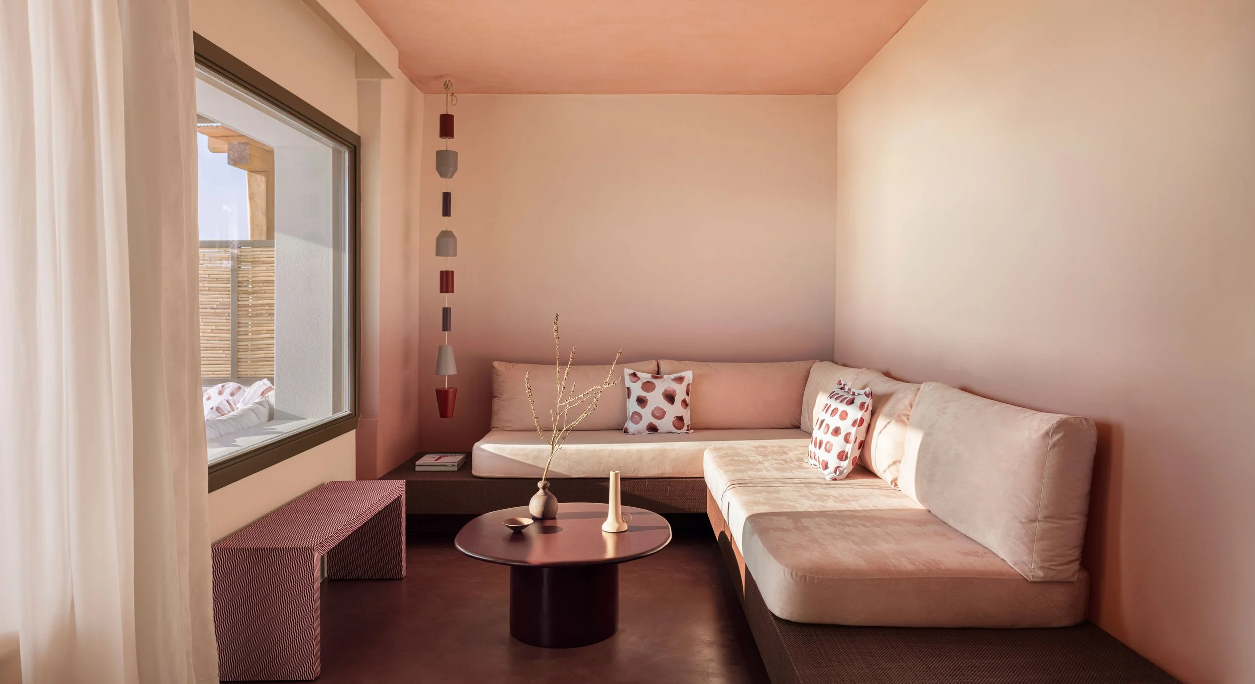 Cozy living room with a large L-shaped beige sofa, decorative pillows with abstract polka dot patterns, a dark round coffee table with vases, a window with sheer curtains, and modern hanging pendant lights.