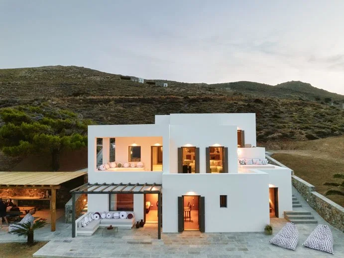 Modern white villa with multiple levels and large windows, surrounded by a hillside landscape, outdoor seating, and lounge chairs.