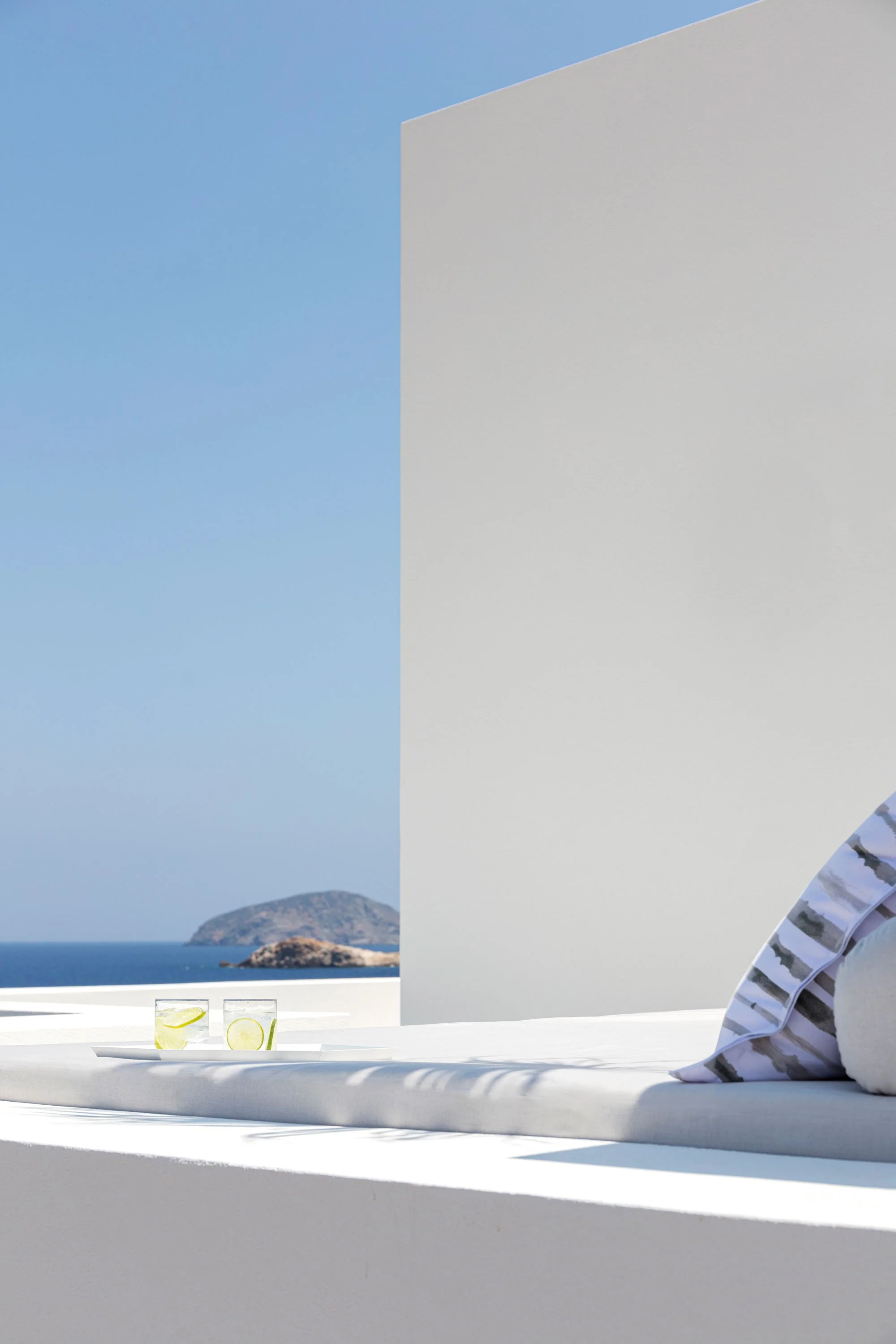 A minimalist outdoor setting with a white bed or lounge area, two glasses of water with lemon slices on a tray, and a view of the ocean and distant islands under a clear blue sky.