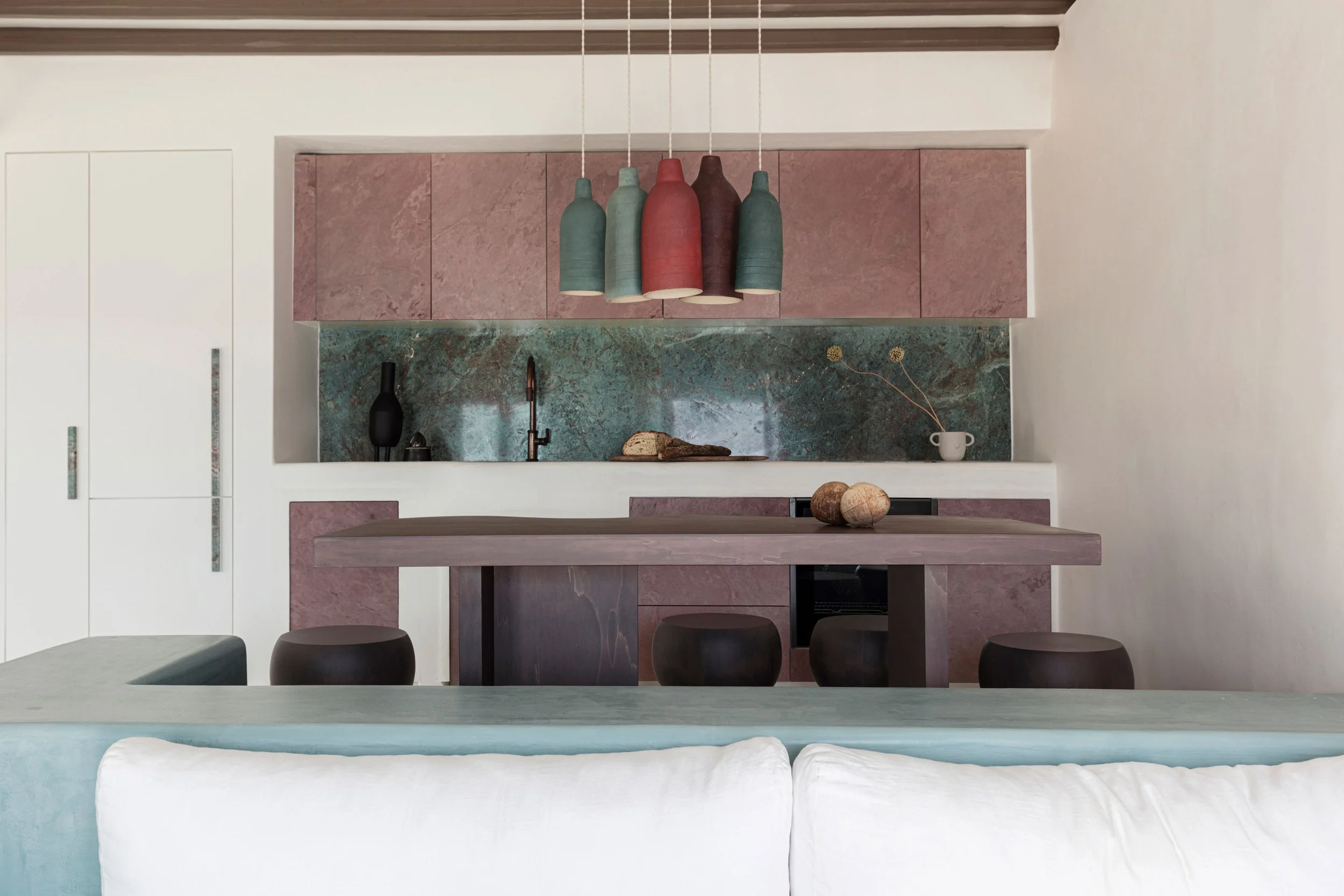 Modern kitchen with pink and green marble backsplash, pink upper cabinets, a wooden dining table, brown stools, and colorful pendant lights.