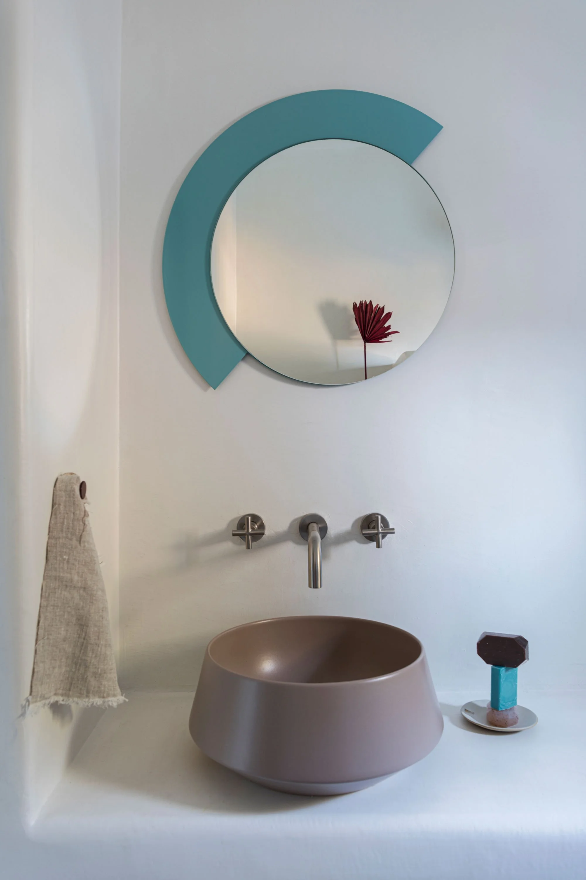 A minimalist bathroom sink area with a round, brown vessel sink, modern wall-mounted faucet, round mirror with a blue semicircular frame, a beige hand towel on a hook, and a small decorative sculpture on a white ledge.