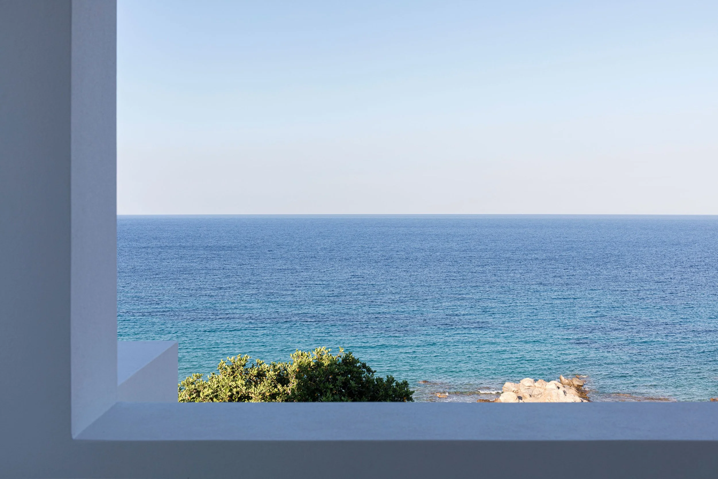 View of the ocean through a window with a white frame, showing blue water, rocks, green foliage, and a clear sky.
