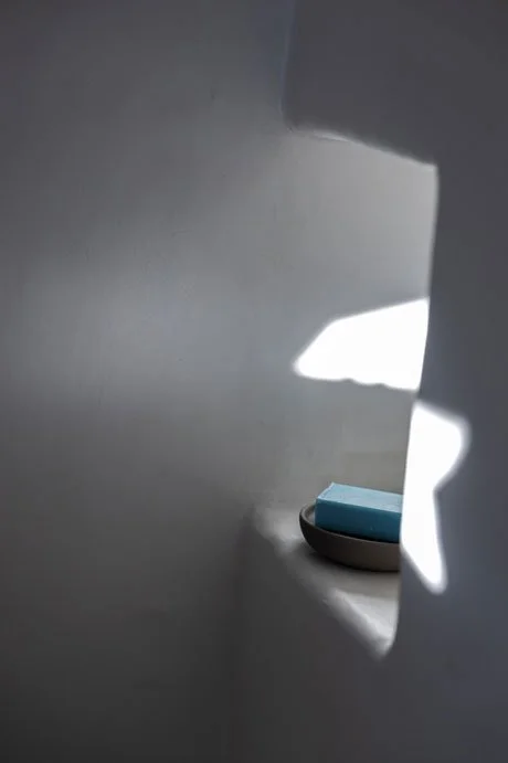 A window with sunlight casting shadow patterns on a wall, with a black dish holding a light blue object near the window.
