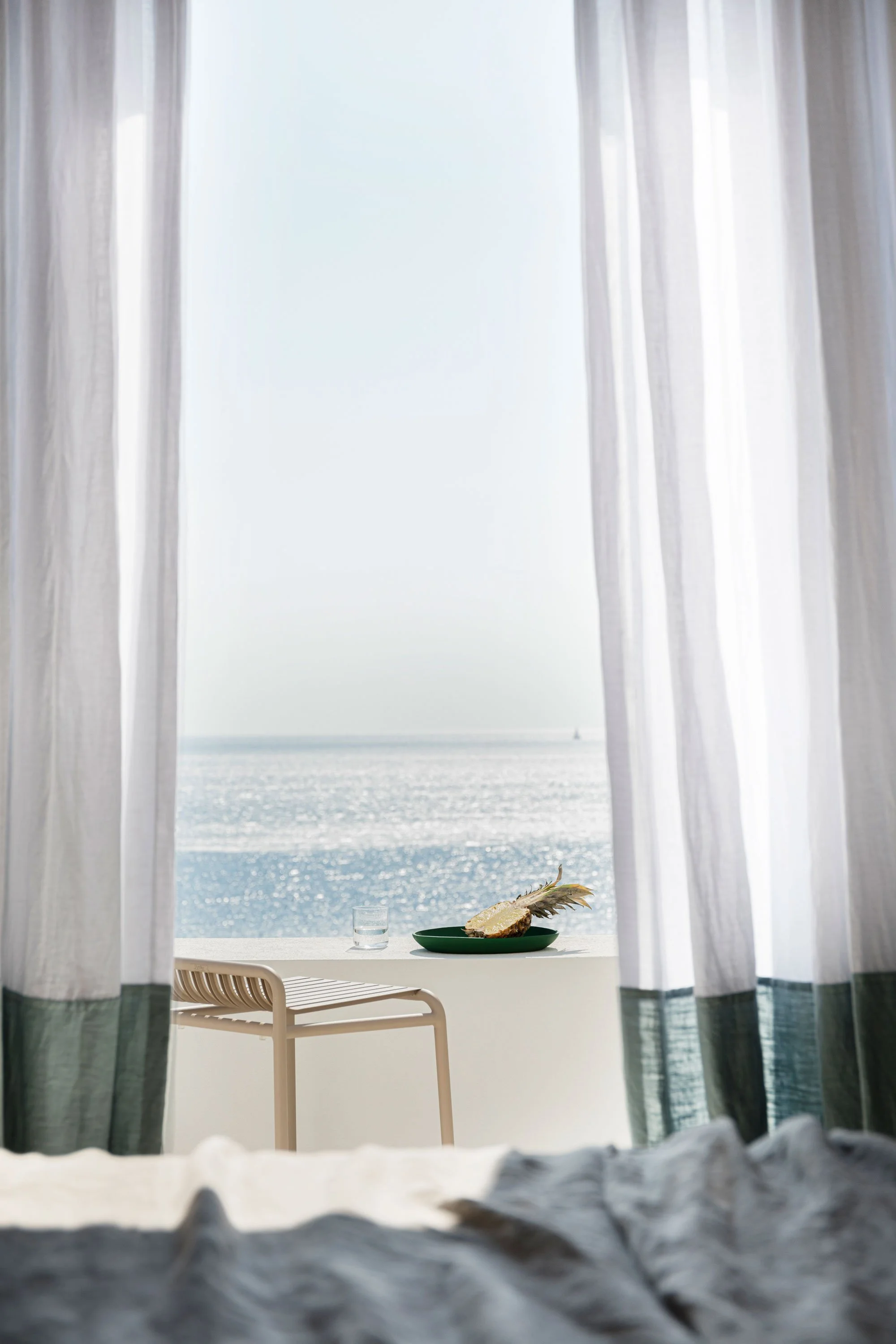 A seaside view through open white curtains, with a white bed in the foreground, a plate with a fish and a feather, a glass of water, and a balcony overlooking the ocean.
